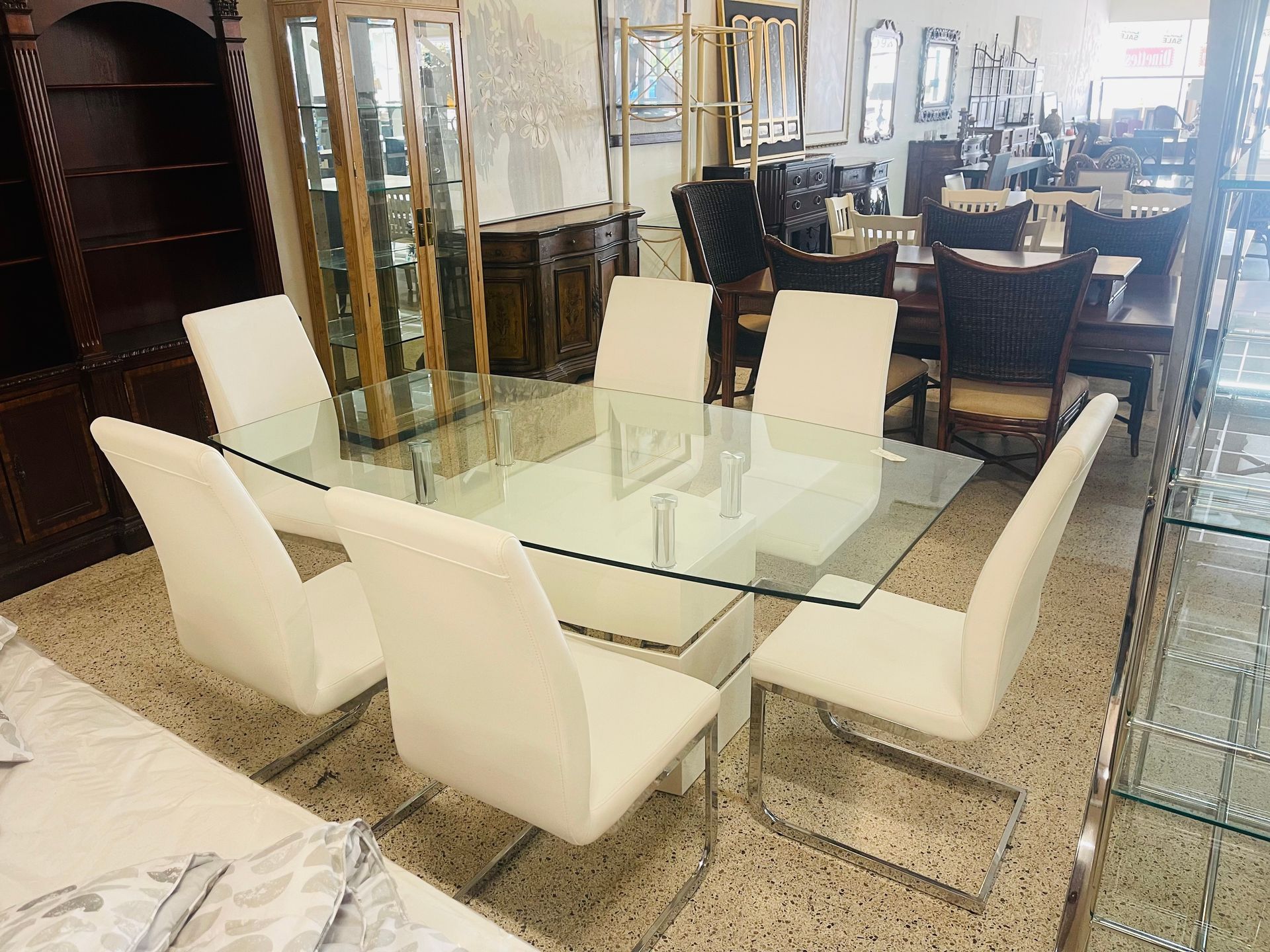 A glass dining table with six cream-colored upholstered chairs on a speckled floor in a furniture store showroom.