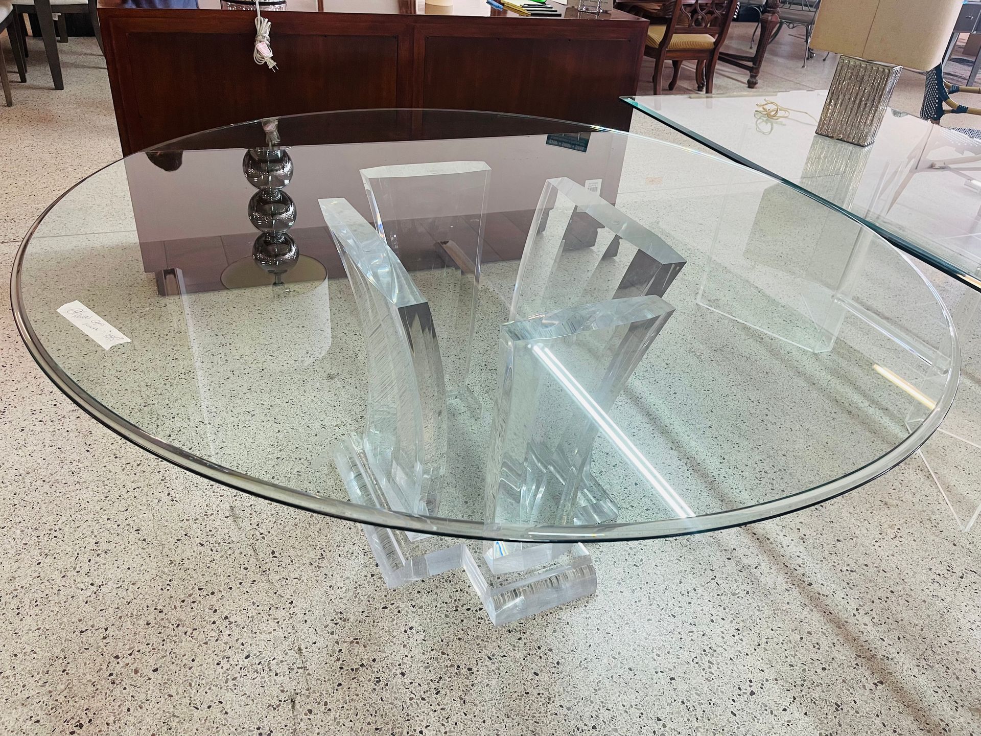 A round, clear glass dining table with a thick, sculpted acrylic pedestal base, shown in a furniture showroom.