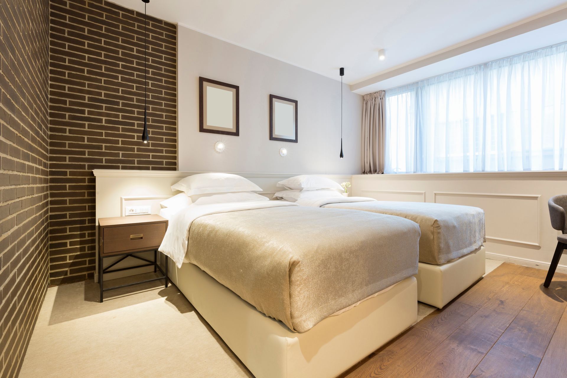 A hotel room with two twin beds, a brown brick accent wall, light-toned bedding, and a large window with sheer curtains.