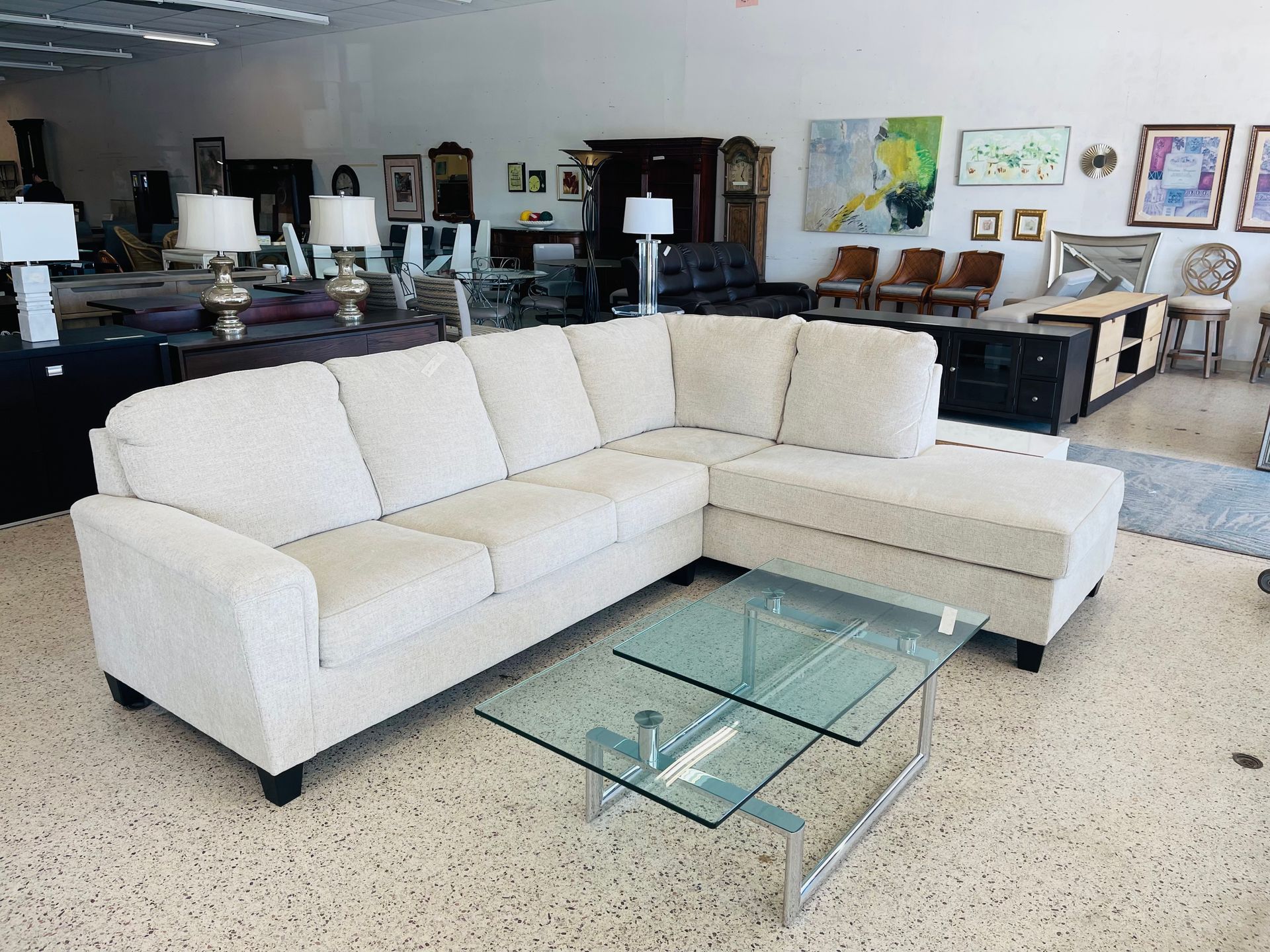 A cream-colored sectional sofa with a glass coffee table in front, displayed in a brightly lit furniture showroom.