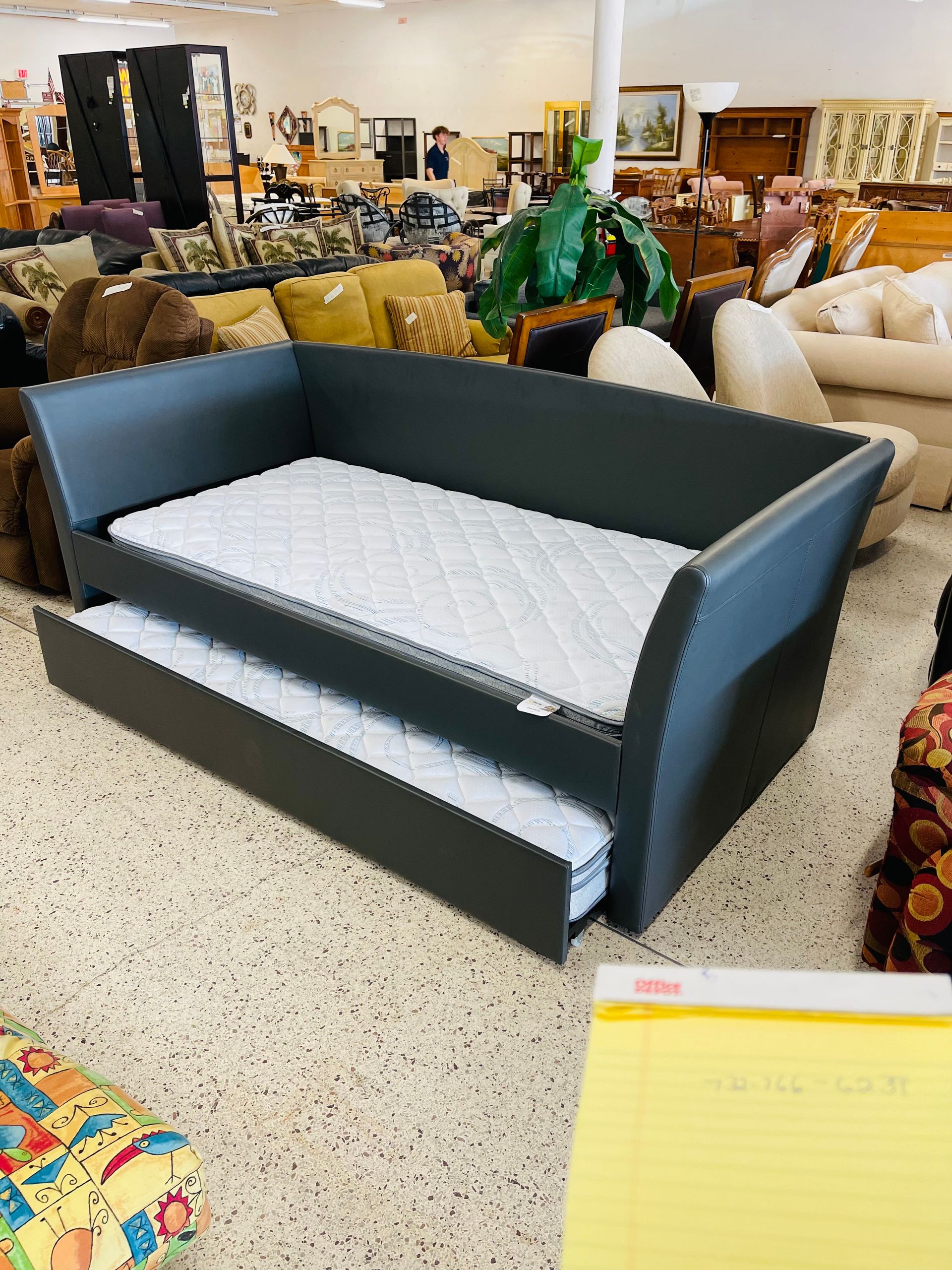 A dark grey upholstered daybed with a pull-out trundle mattress sitting in a furniture store showroom.