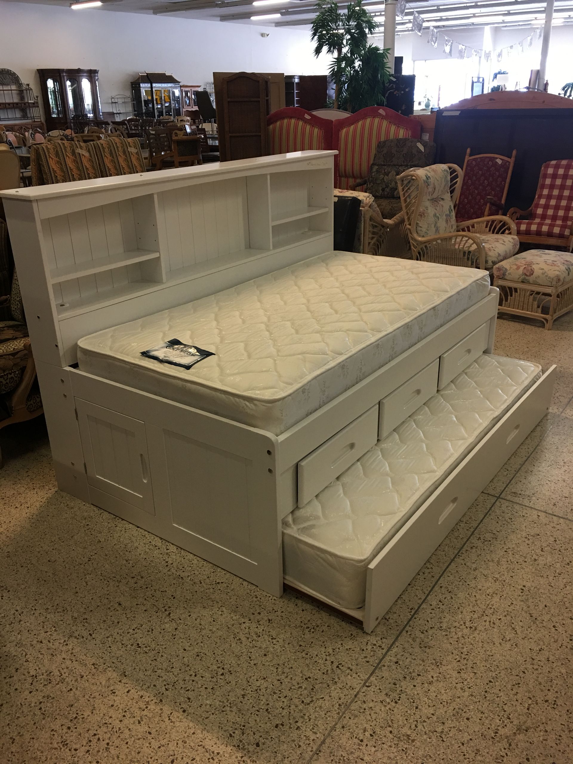 A white storage bed frame with a headboard containing shelves and a pull-out trundle bed, displayed in a furniture store.