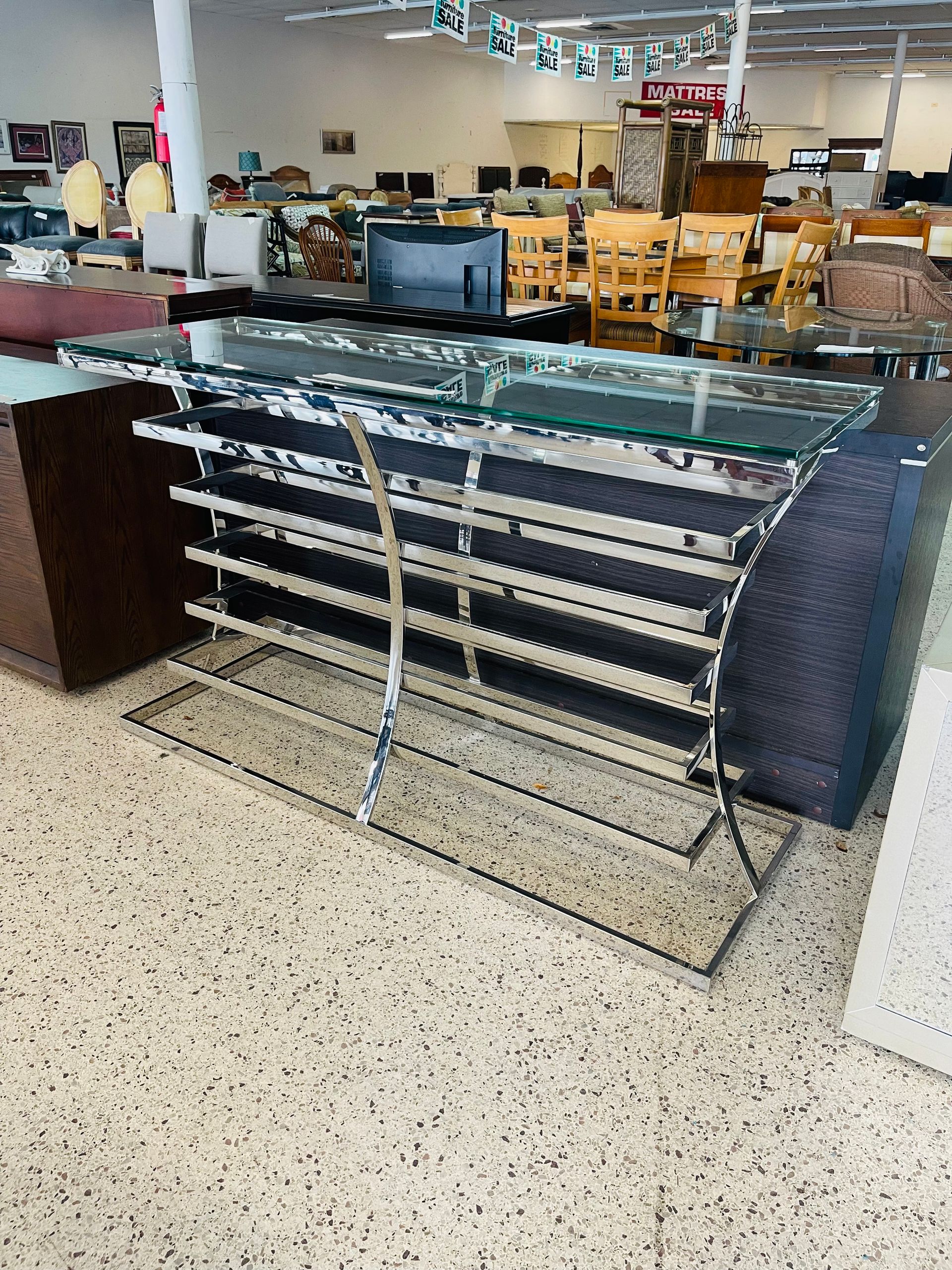 A glass-topped chrome console table with tiered shelving sits on a speckled floor inside a furniture store showroom.