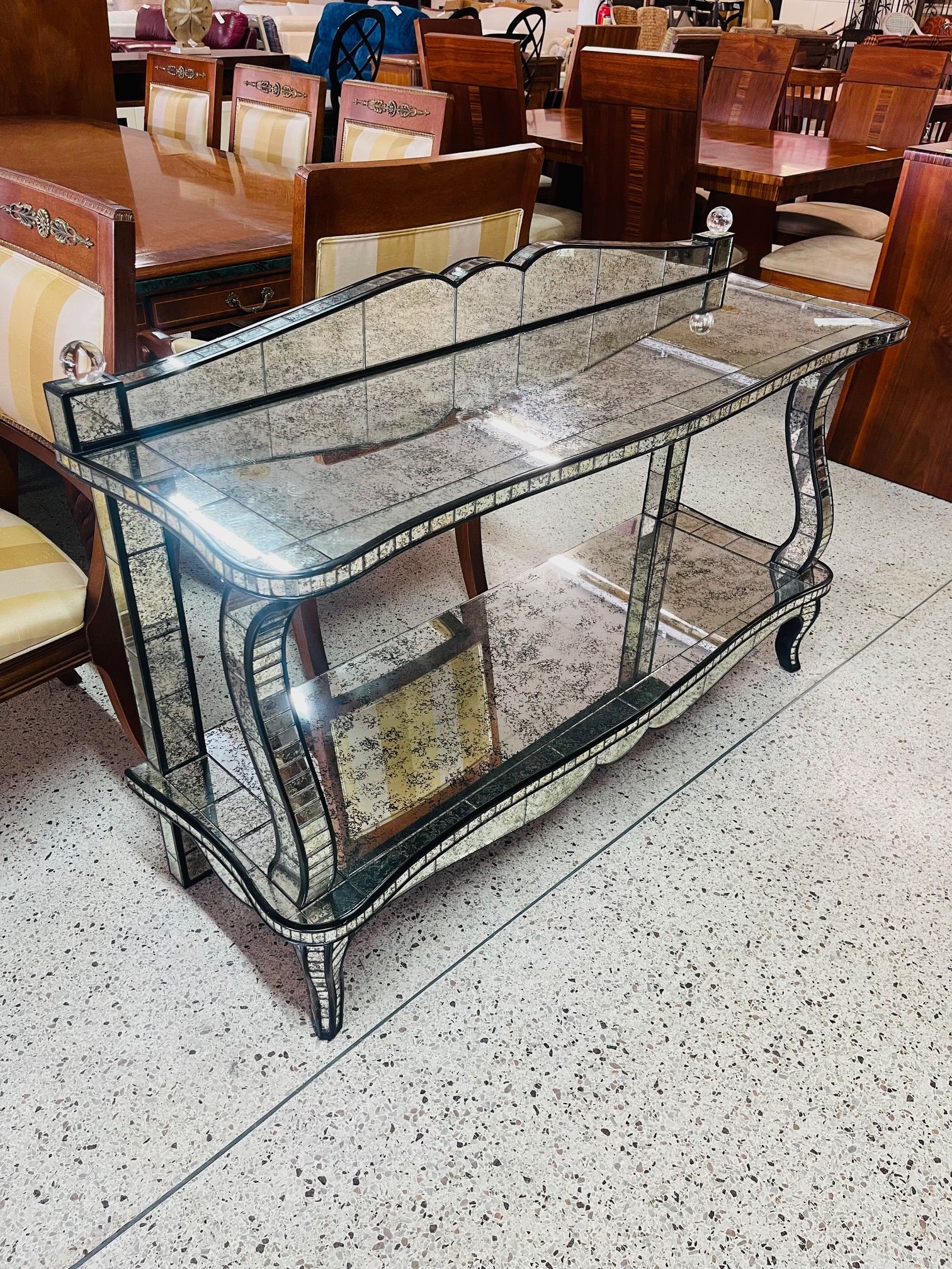 A mirrored console table with a shelf stands in a furniture store showroom with wooden chairs in the background.