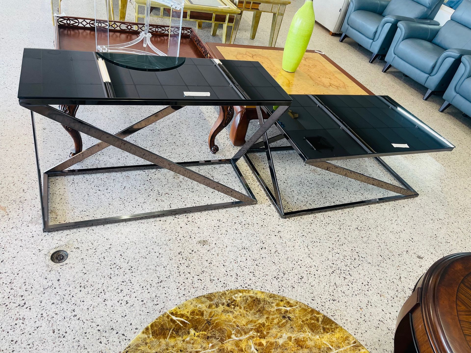 Two rectangular black side tables with geometric metal bases sit on a speckled floor near furniture in a showroom.