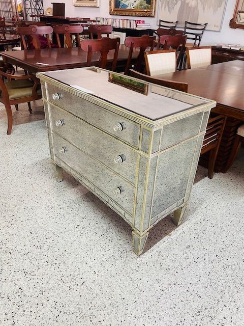 A mirrored three-drawer chest with glass knobs, standing on a white speckled floor in a furniture store.