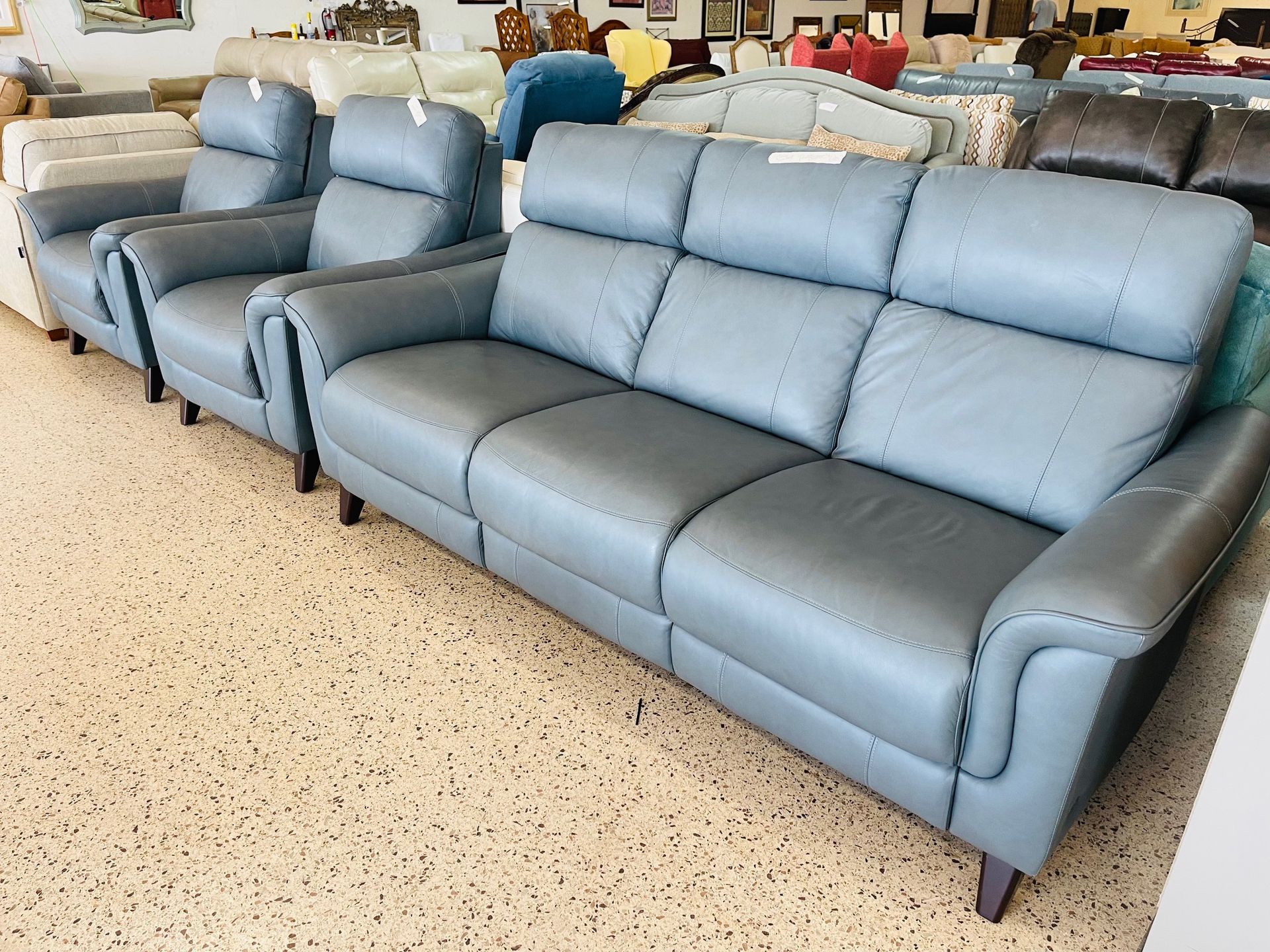 A light blue leather three-seat sofa sits in a furniture store showroom next to two matching armchairs.