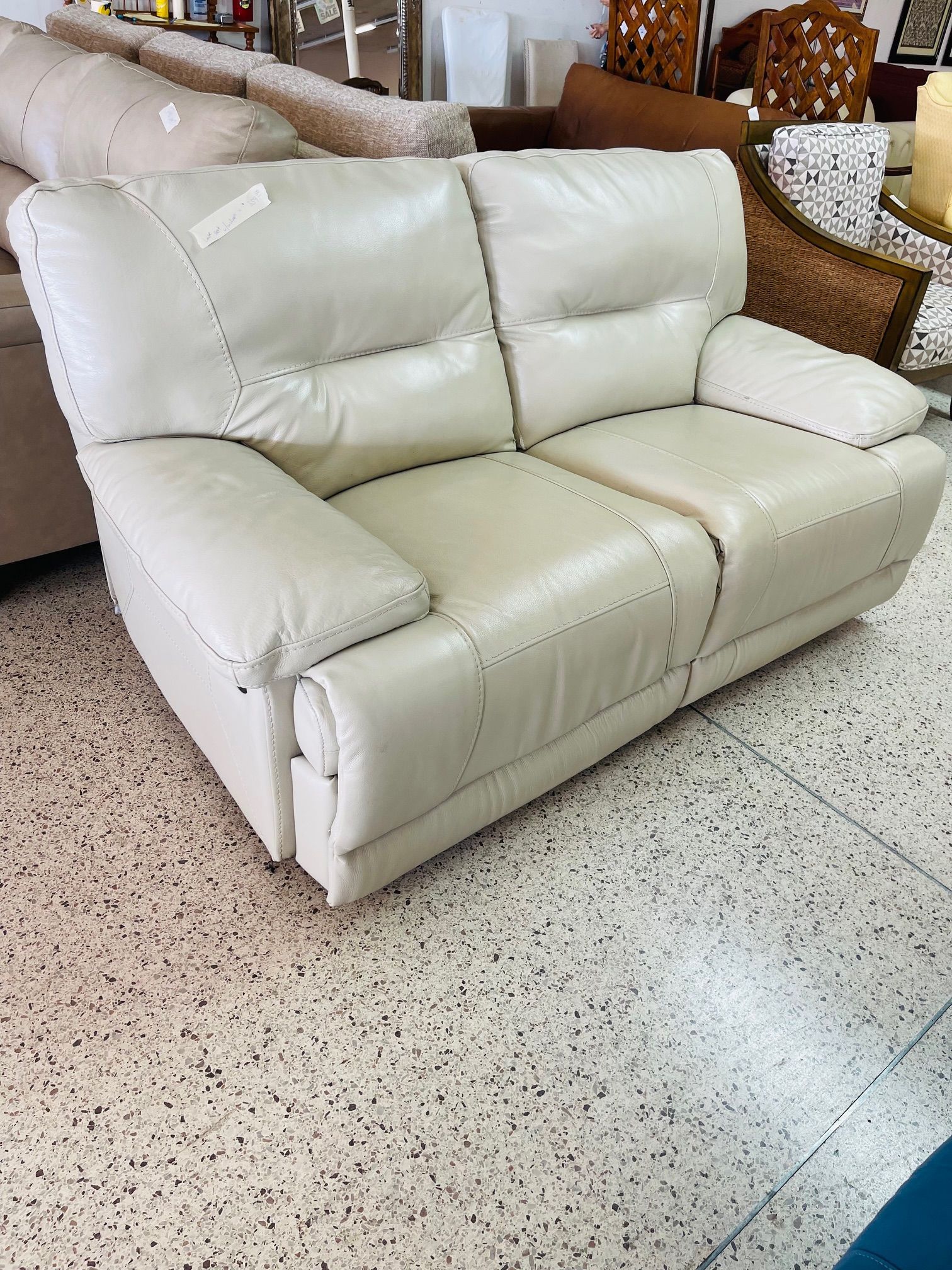 A white, two-seat leather reclining sofa sits on a speckled tile floor in a furniture store.