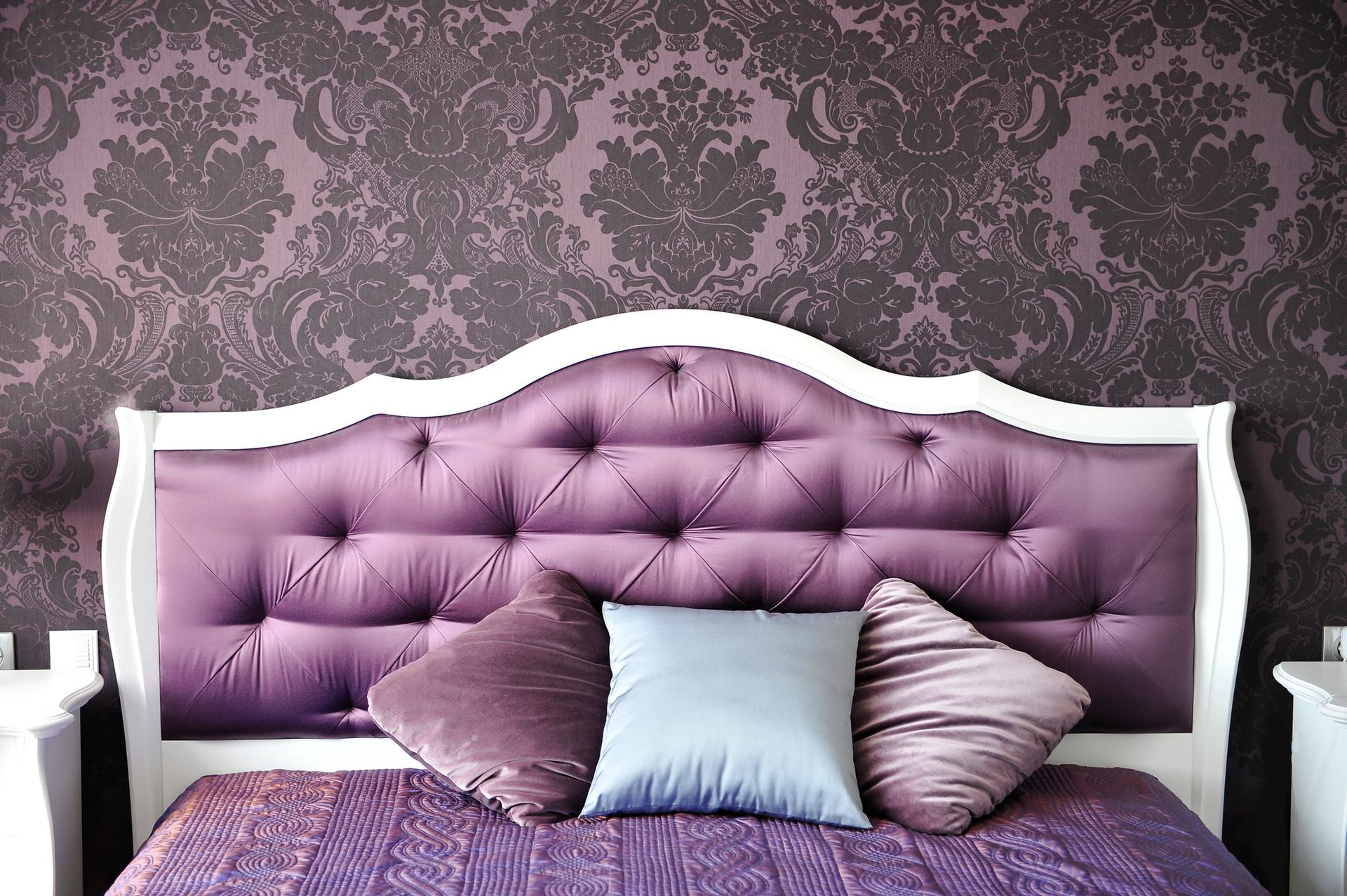 A tufted purple headboard with white trim against patterned dark wallpaper, styled with three plush pillows on a bed.