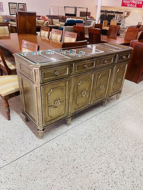 A vintage-style buffet or sideboard with a distressed gold finish, featuring drawers and cabinets, in a furniture store.