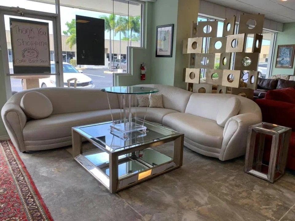 A beige curved sectional sofa sits in a store showroom, featuring a glass-topped end table and a mirrored coffee table.