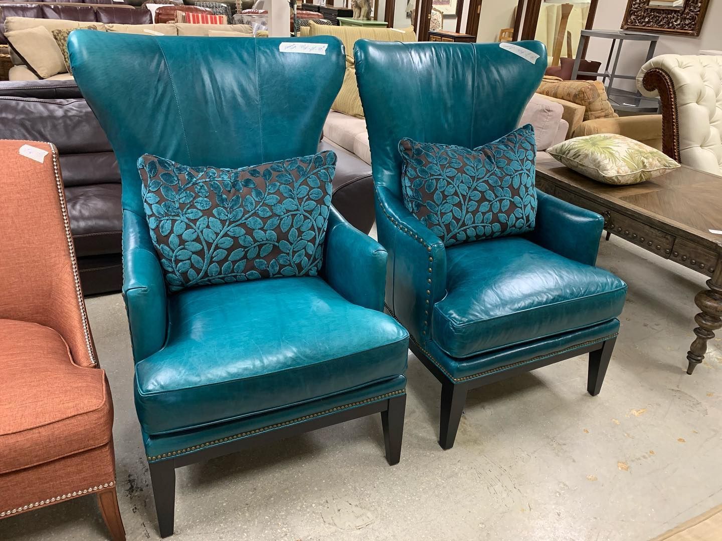 Two vibrant teal leather wingback chairs with patterned accent pillows on display in a furniture store.