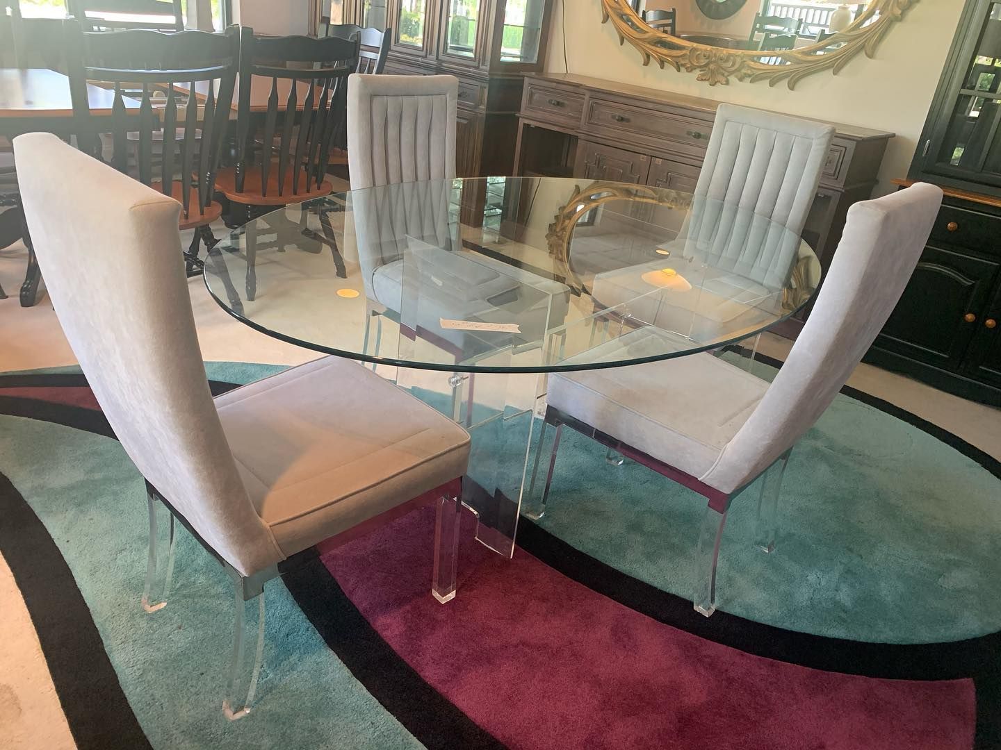 A glass-topped dining table with four light-colored upholstered chairs on a vibrant, multi-colored circular rug.
