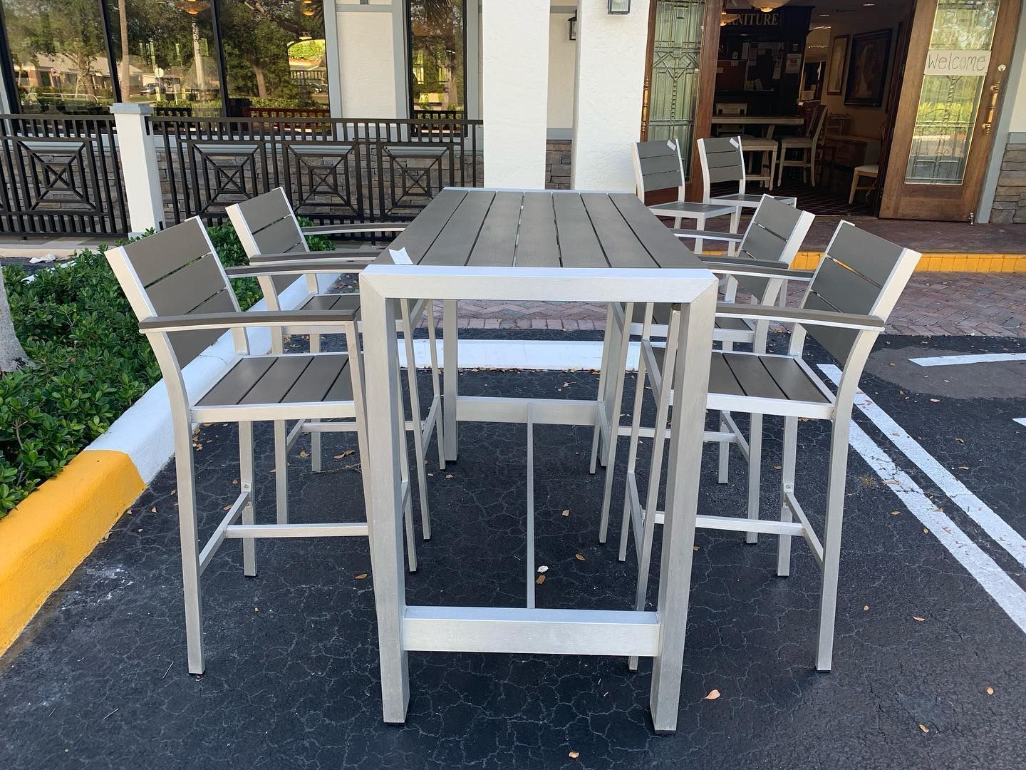 A tall outdoor rectangular table with four matching high-back chairs on asphalt near a building entrance.