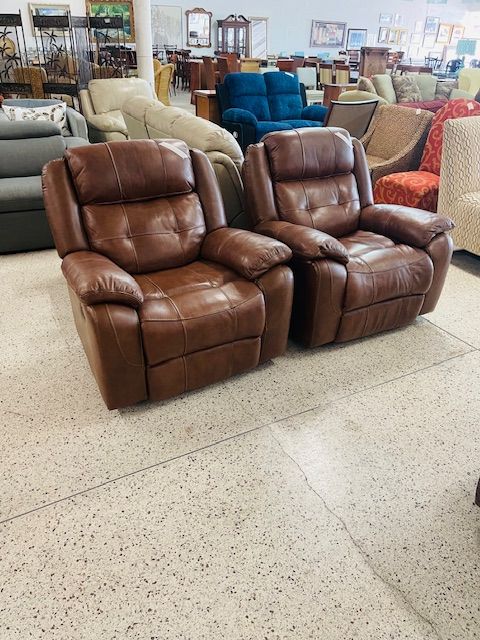 Two brown leather recliners side-by-side in a furniture store showroom with other chairs in the background.