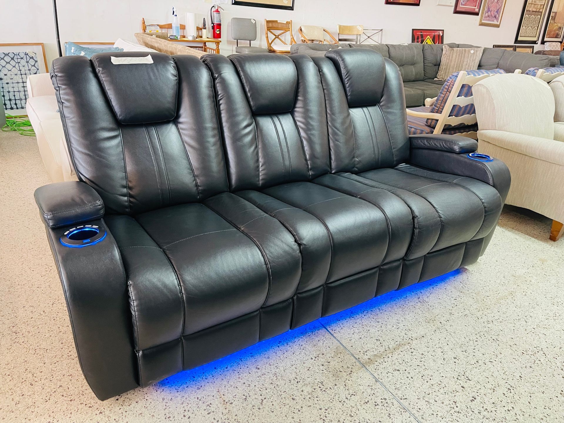 A black three-seat reclining sofa with integrated blue-lit cup holders and base lighting in a furniture showroom.