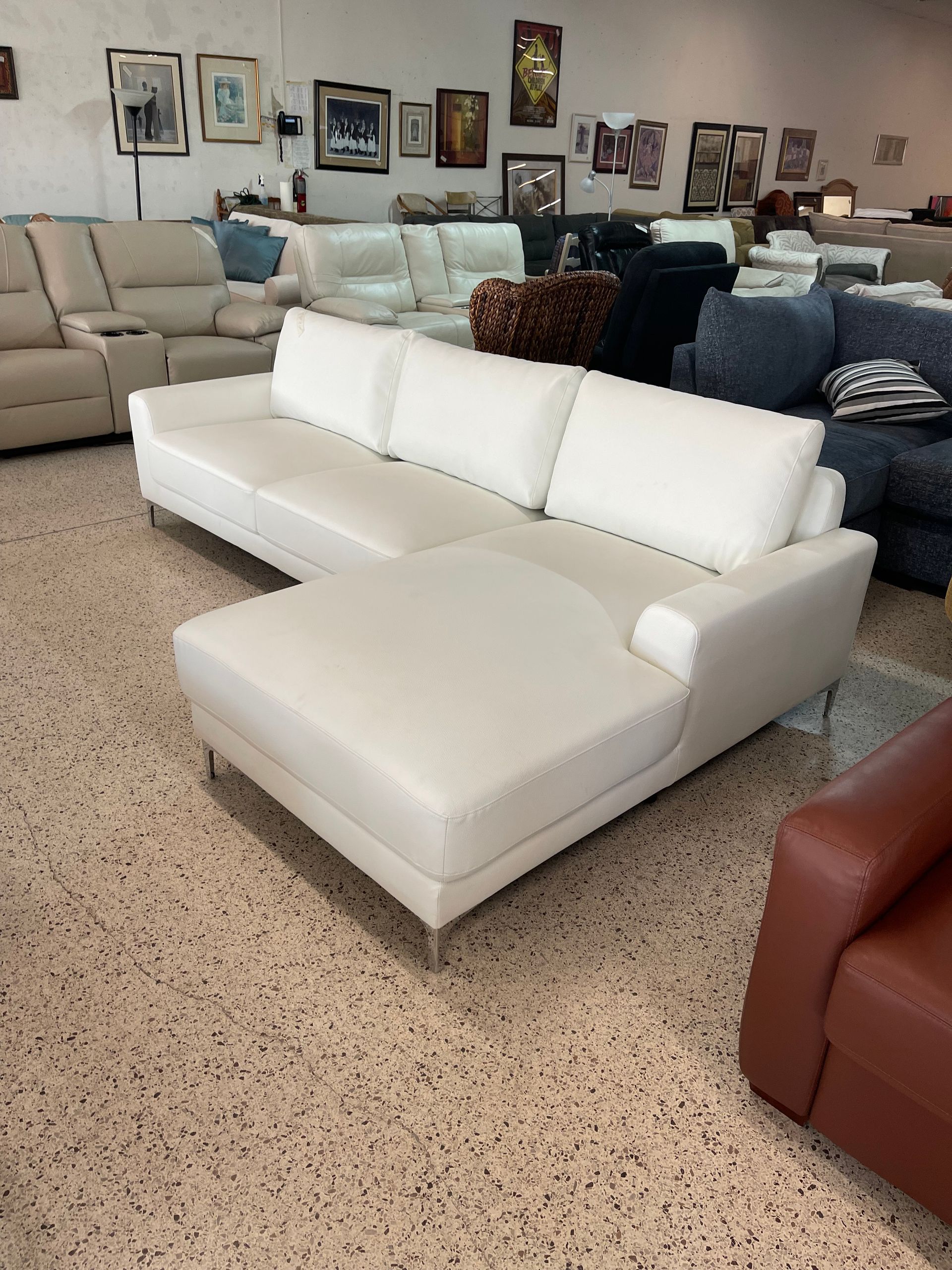 A modern white sectional sofa with a chaise lounge sits on a speckled floor in a furniture showroom.