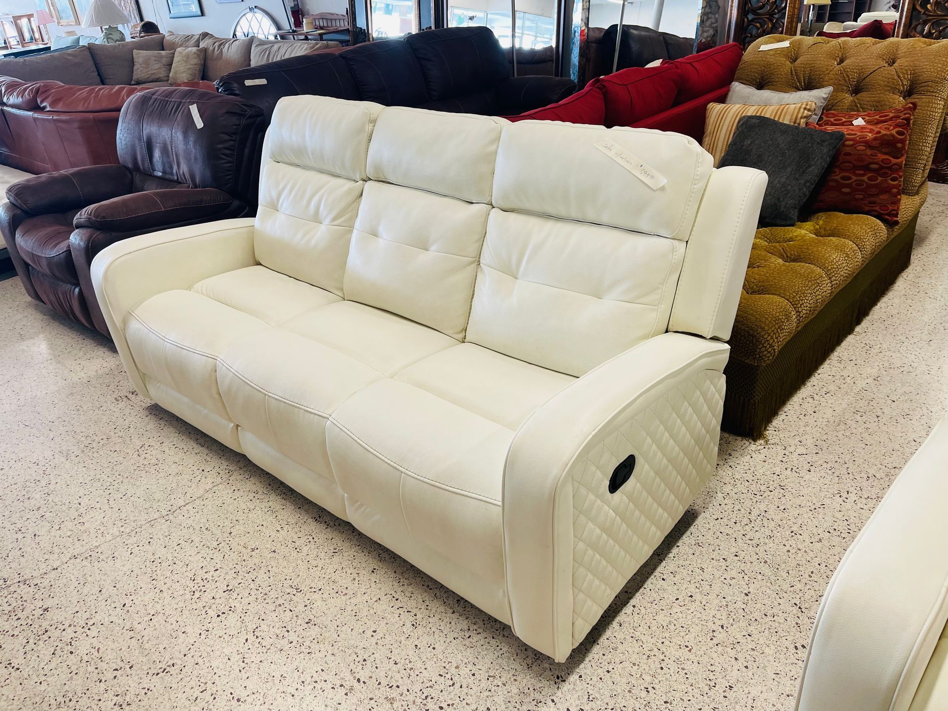 A white, three-cushion leather recliner sofa displayed in a furniture showroom next to other sofas.