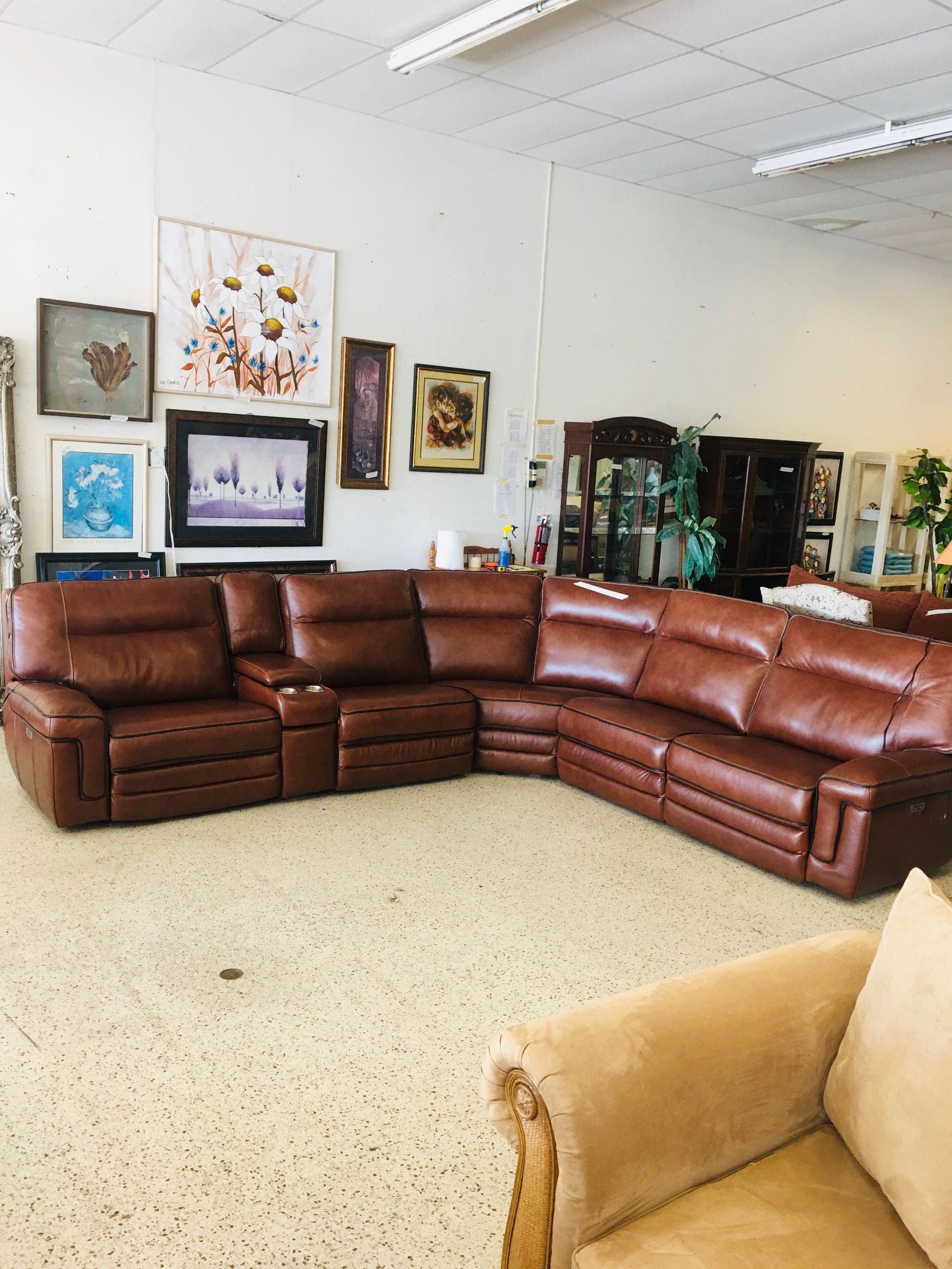 A large, brown leather sectional sofa sits in a brightly lit furniture store, with framed art displayed on the wall.