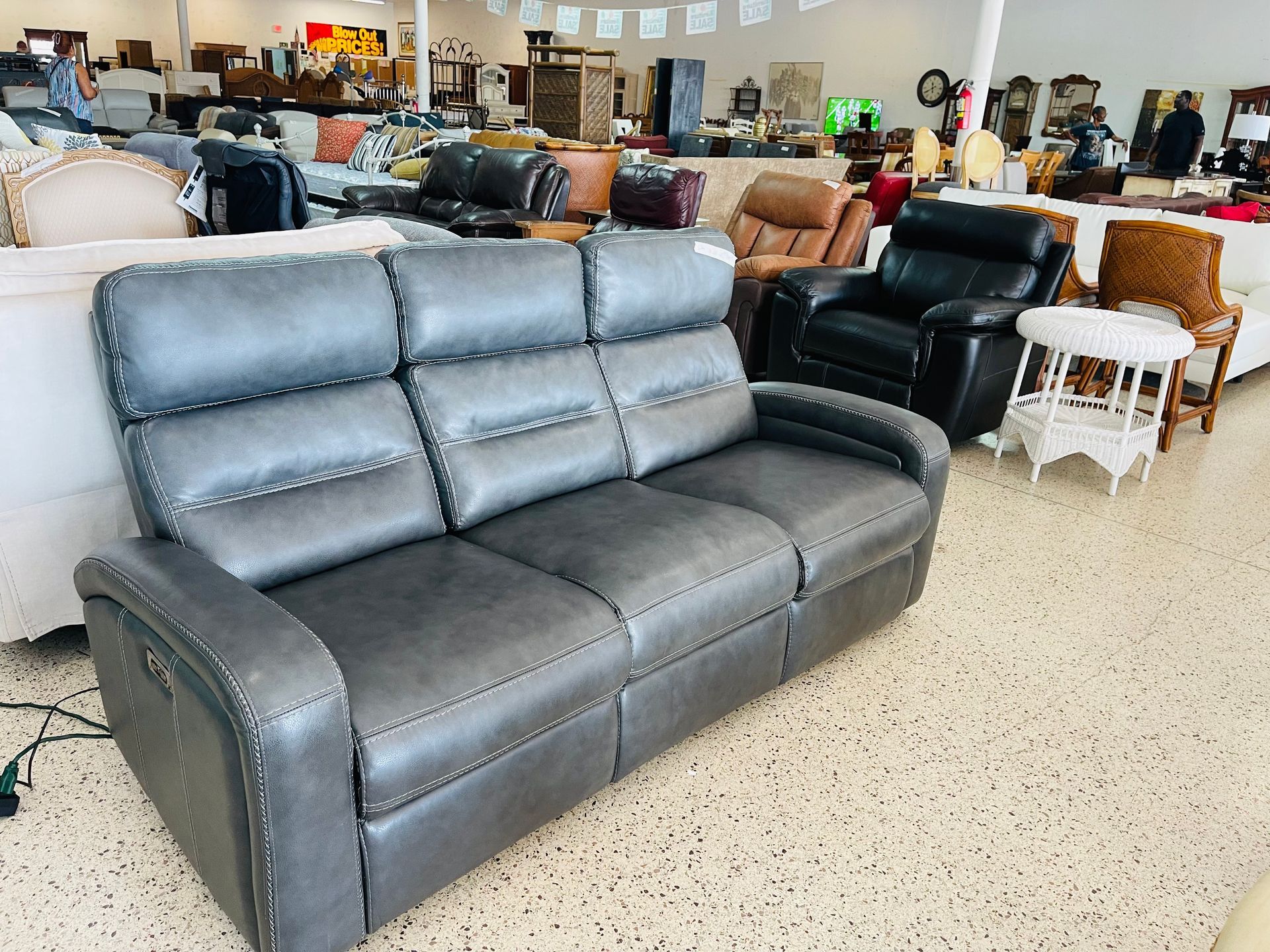 A gray, three-cushion reclining sofa with nailhead trim sits in a well-lit furniture showroom.