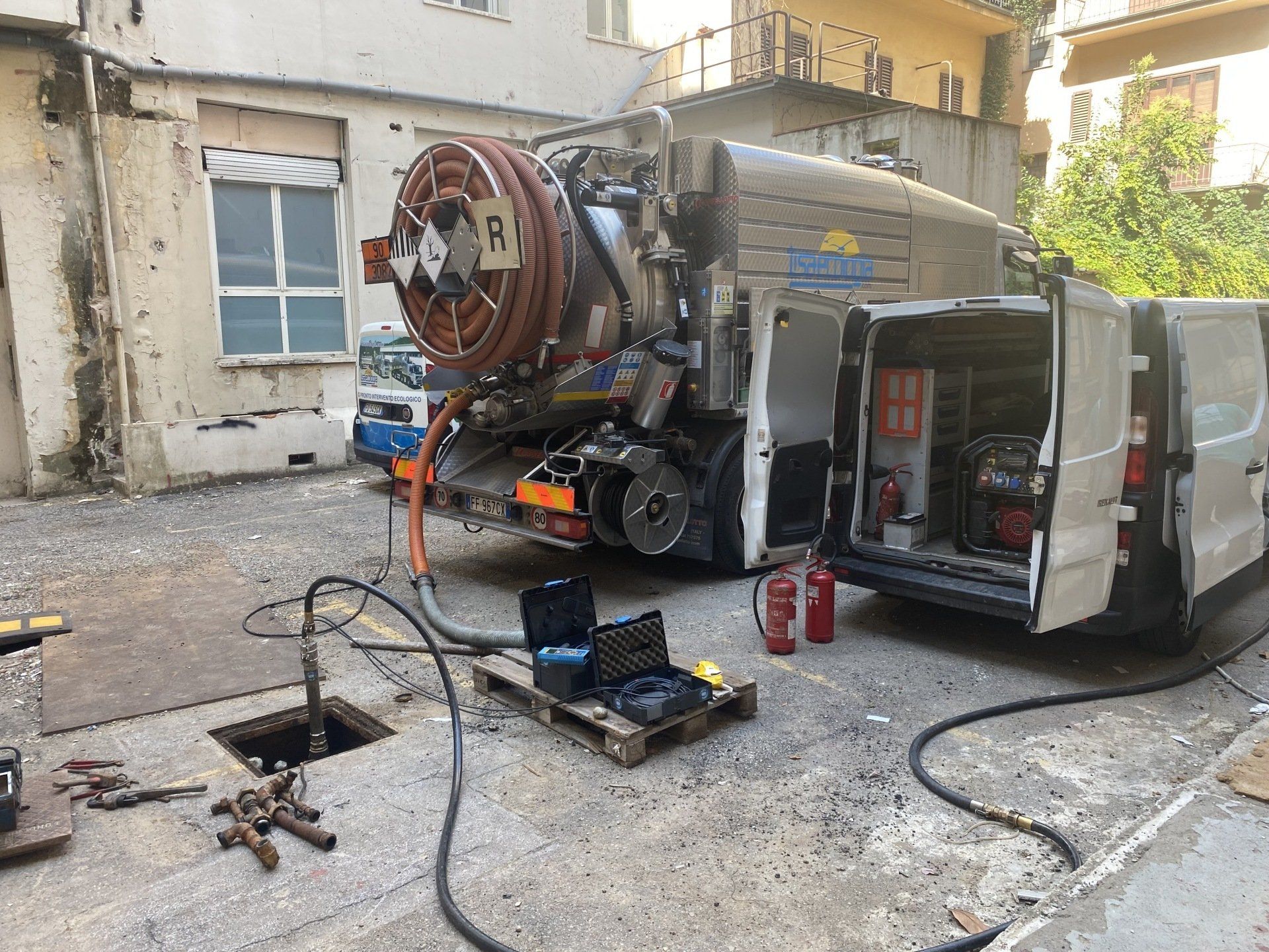 Un camion per la nettezza urbana con le porte aperte, i tubi flessibili collegati a un tombino aperto e un furgone accanto.