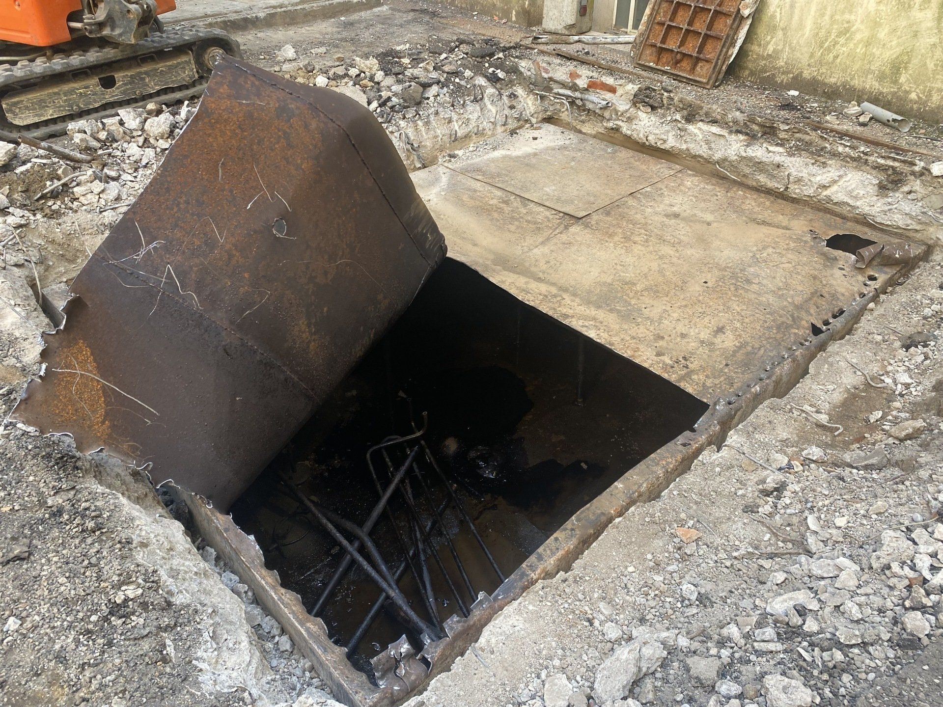 Tombino aperto con coperchio di metallo arrugginito, circondato da detriti.
