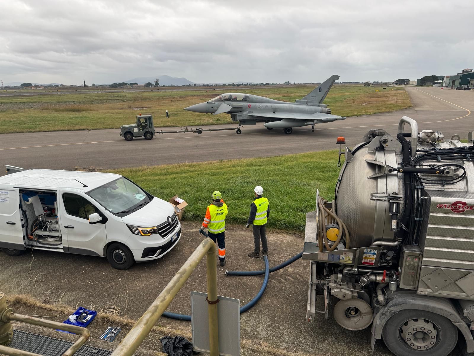 Operai che riforniscono di carburante un aereo da caccia su un aeroporto, con un furgone e un camion per la manutenzione presenti sotto un cielo coperto.