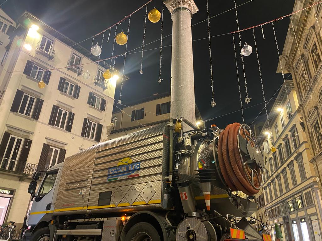 Un grande camion industriale parcheggiato in una strada cittadina di notte. Il camion è argentato e ha un grosso tubo flessibile attaccato. Edifici e luci natalizie sullo sfondo.