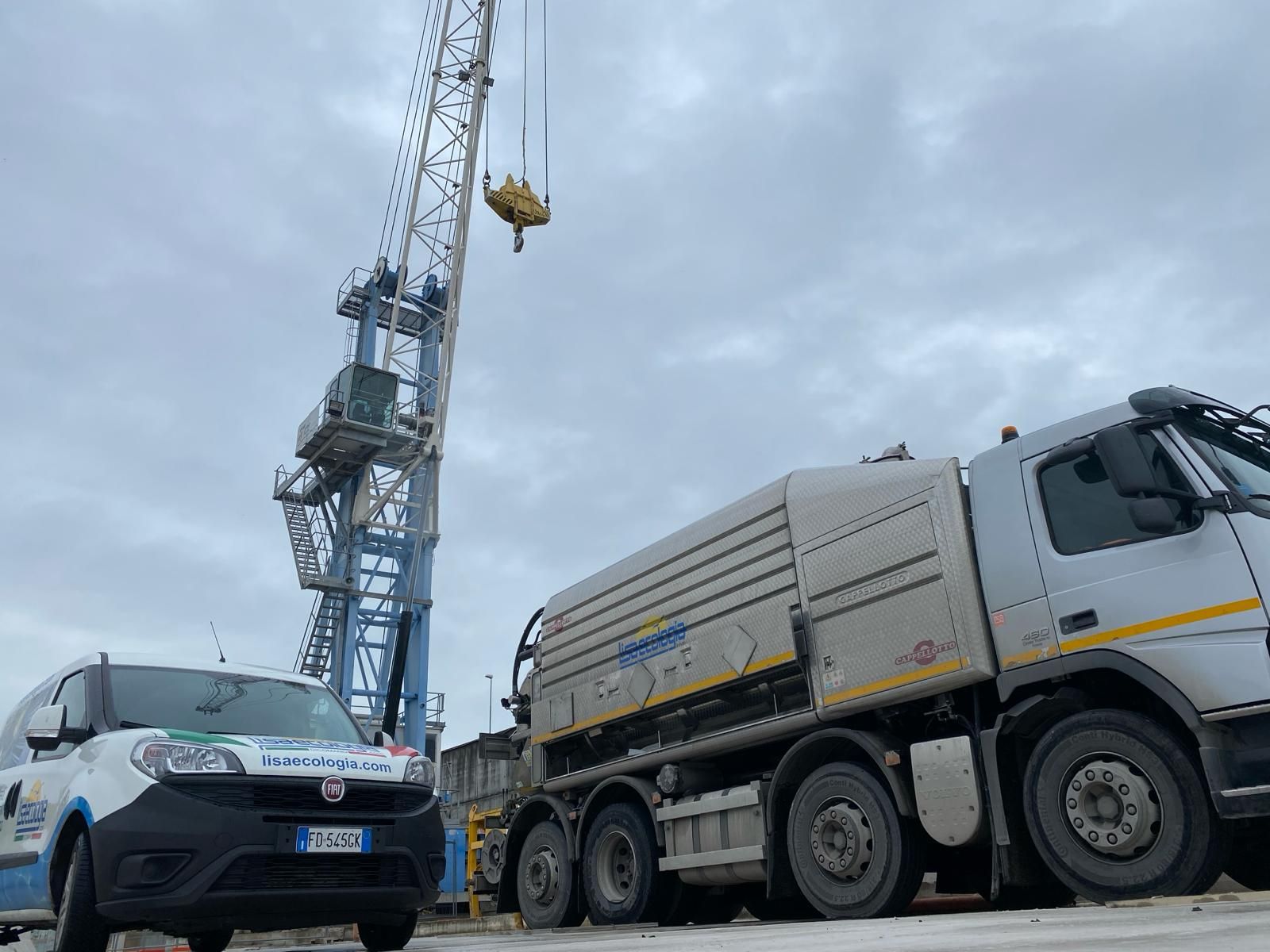 Gru e due camion in una giornata nuvolosa. Un camion è bianco e grigio, l'altro è bianco e blu.