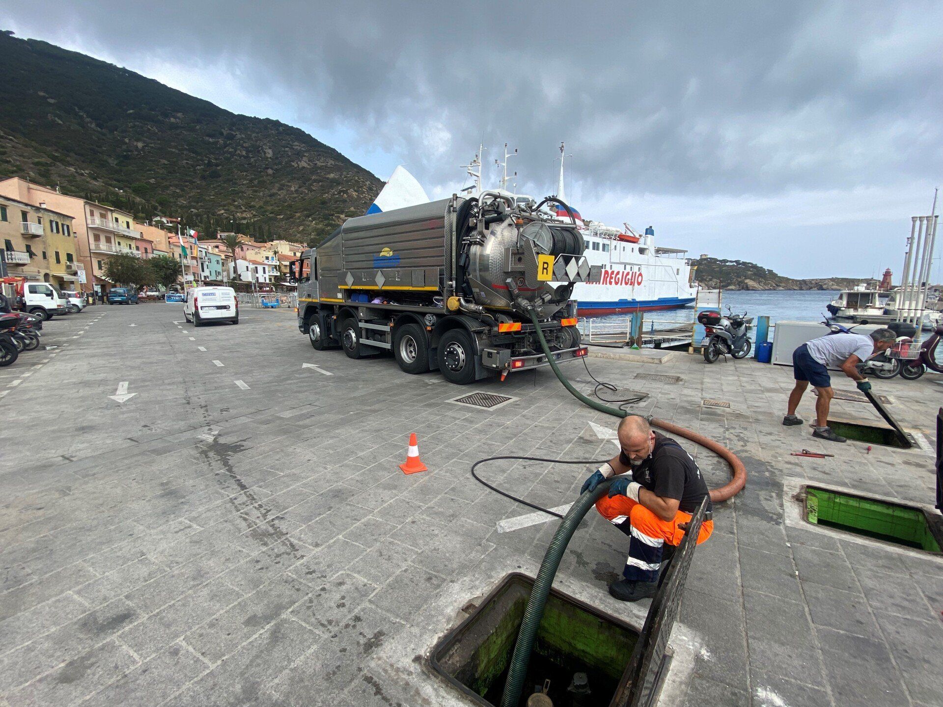 Un camion aspiratutto pulisce una fogna in un porto, un operaio collega un tubo. Sono visibili il traghetto e la città. Cielo nuvoloso.