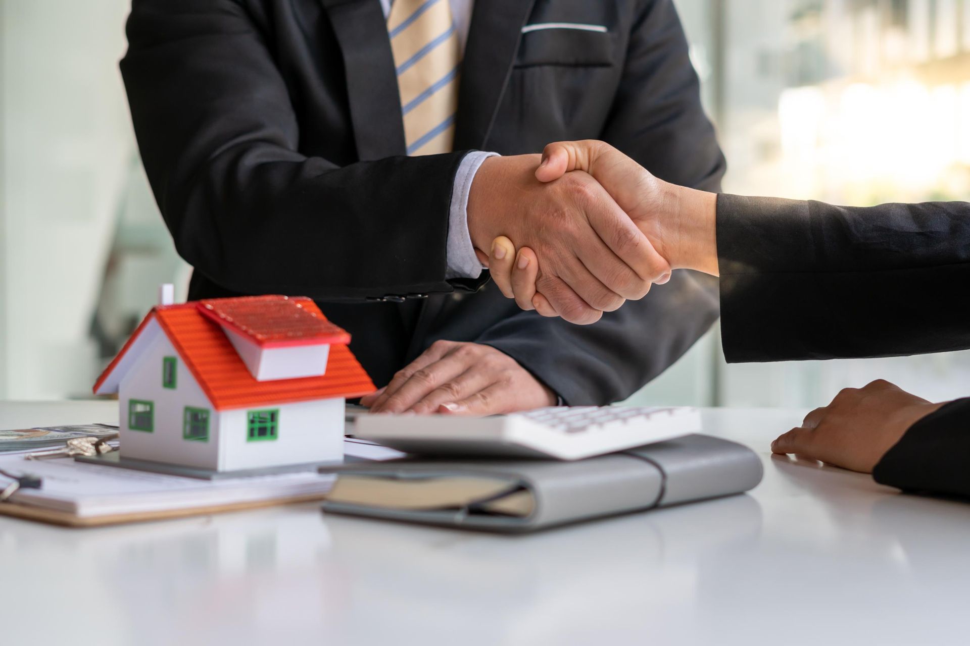 Two people shaking hands over a table with a house model, calculator, and papers, likely a real estate deal.