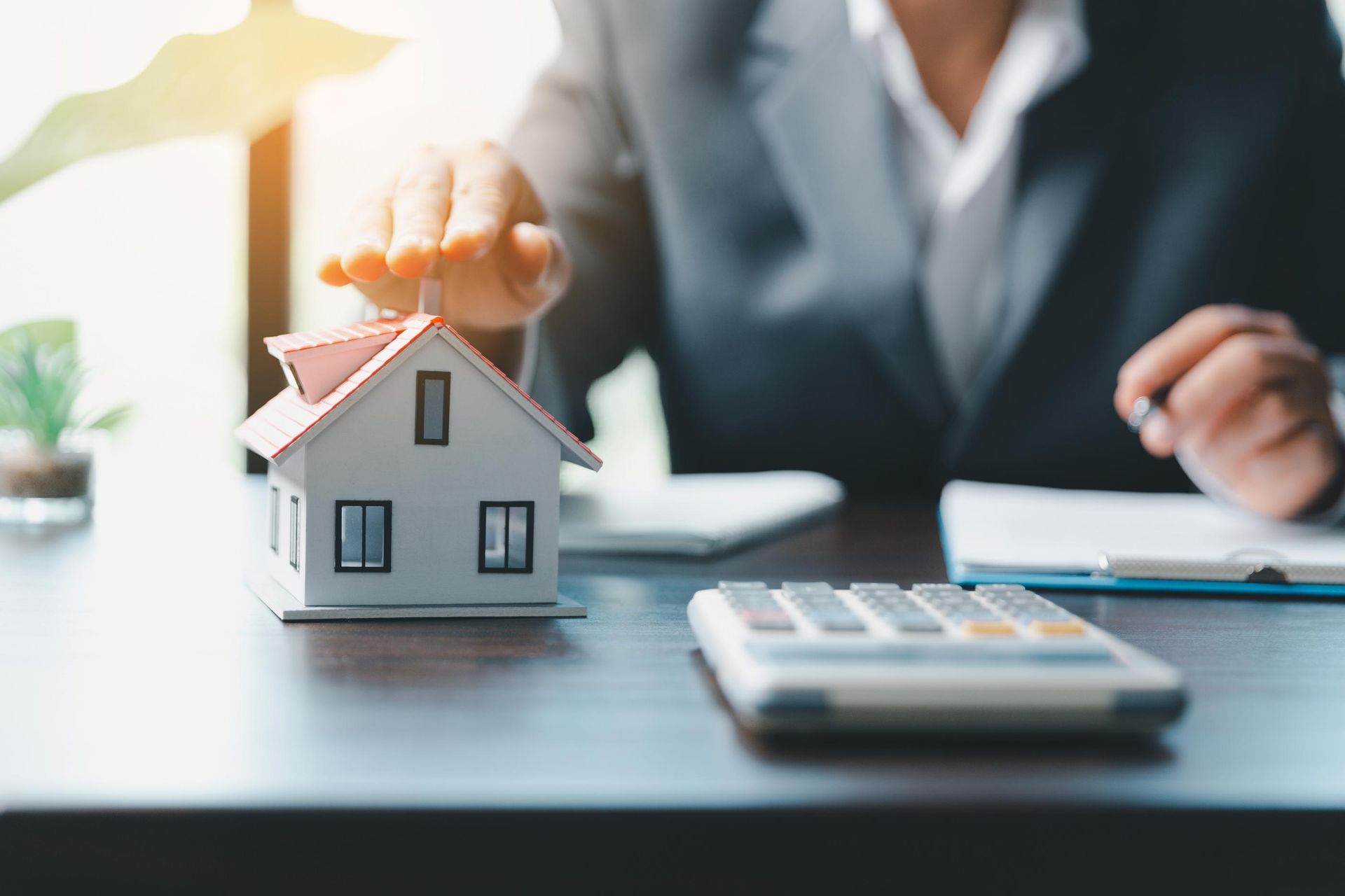 Person's hand protecting a miniature house on a desk with a calculator and documents.