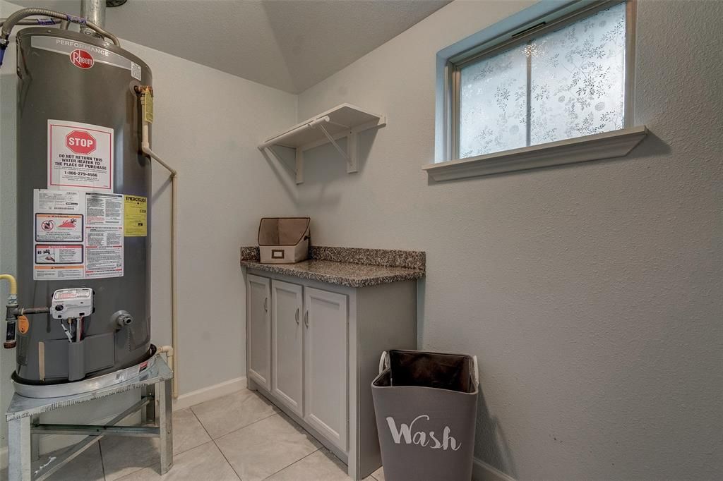 A laundry room with a water heater , cabinets , a trash can and a window.