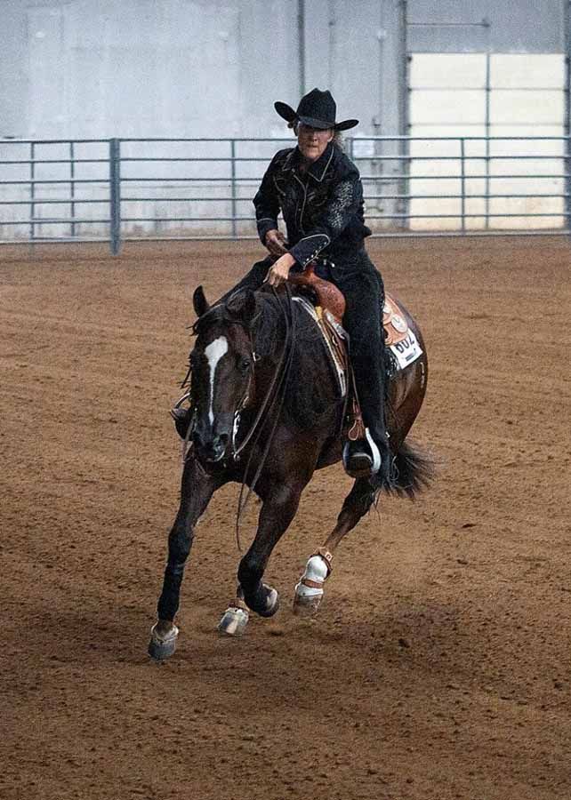 Cowgirl in black riding a dark horse in an indoor arena, turning sharply in the dirt.