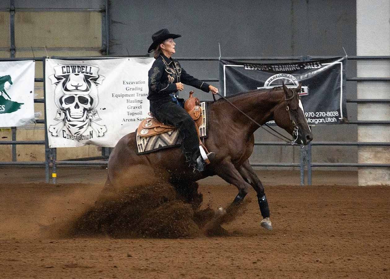 Cowgirl riding a brown horse, skidding through an indoor arena with dirt spraying behind.