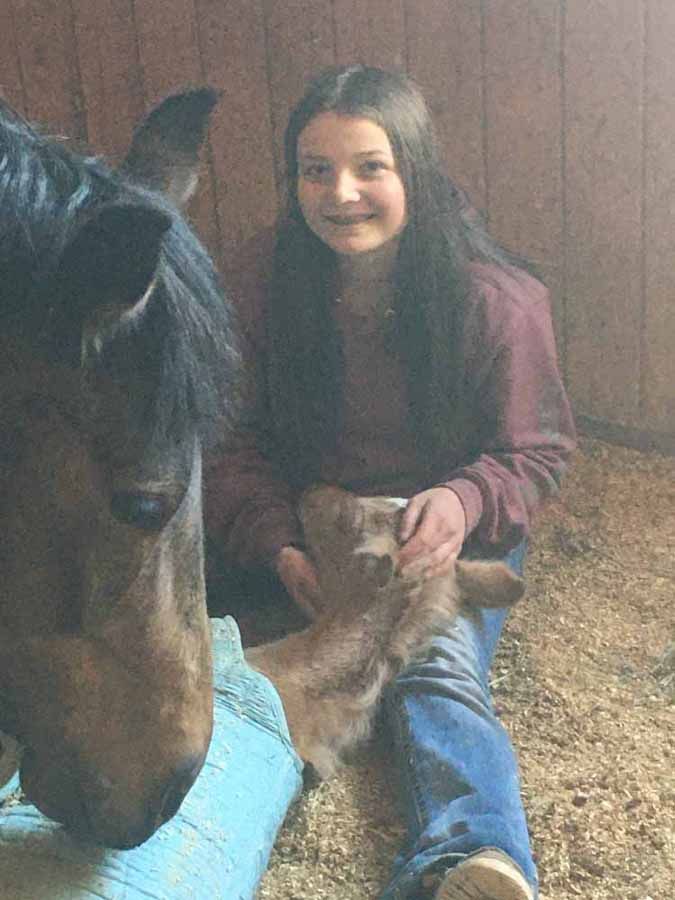 Person with long dark hair smiling beside a horse in a barn, holding its hoof.