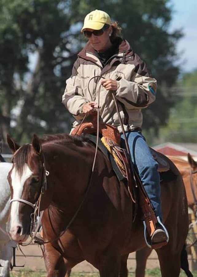 Person riding a brown horse outdoors, wearing a beige jacket and yellow cap.