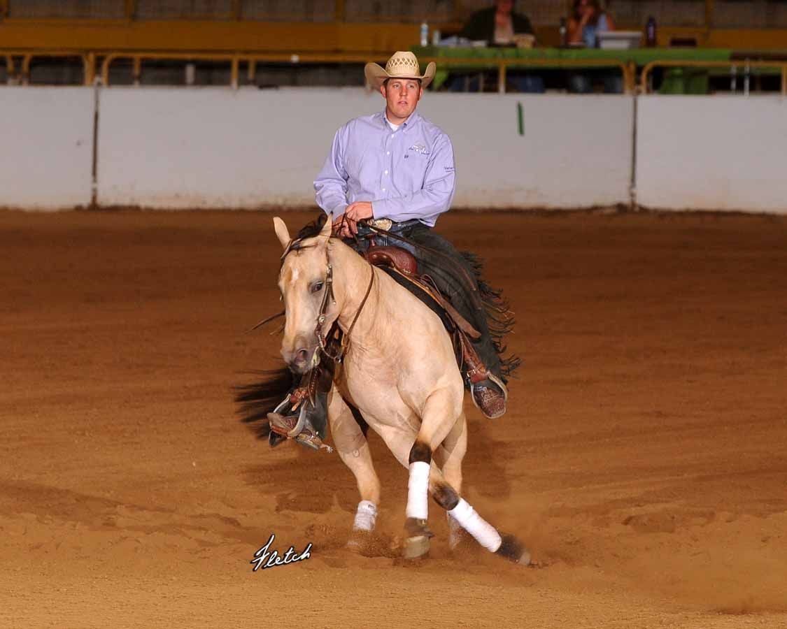 Cowboy riding a light-colored horse in an indoor arena, leaning into a turn.