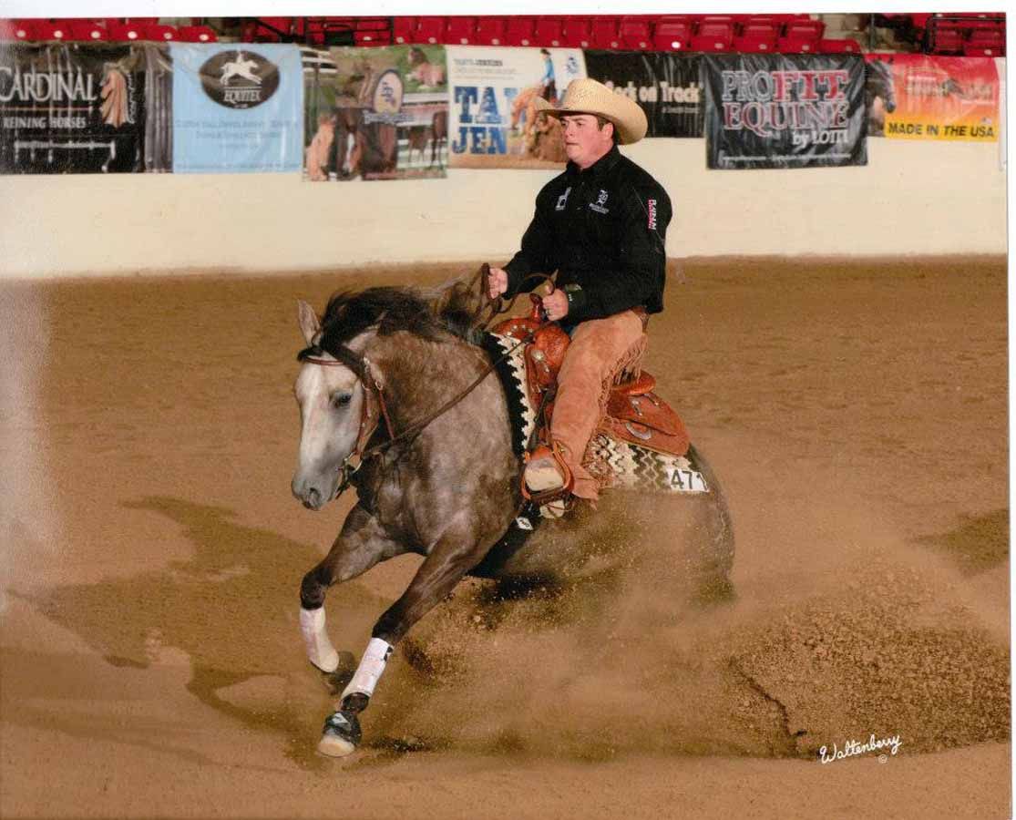 Cowboy riding a gray horse fast around an indoor arena, kicking up dust, with banners on the wall.