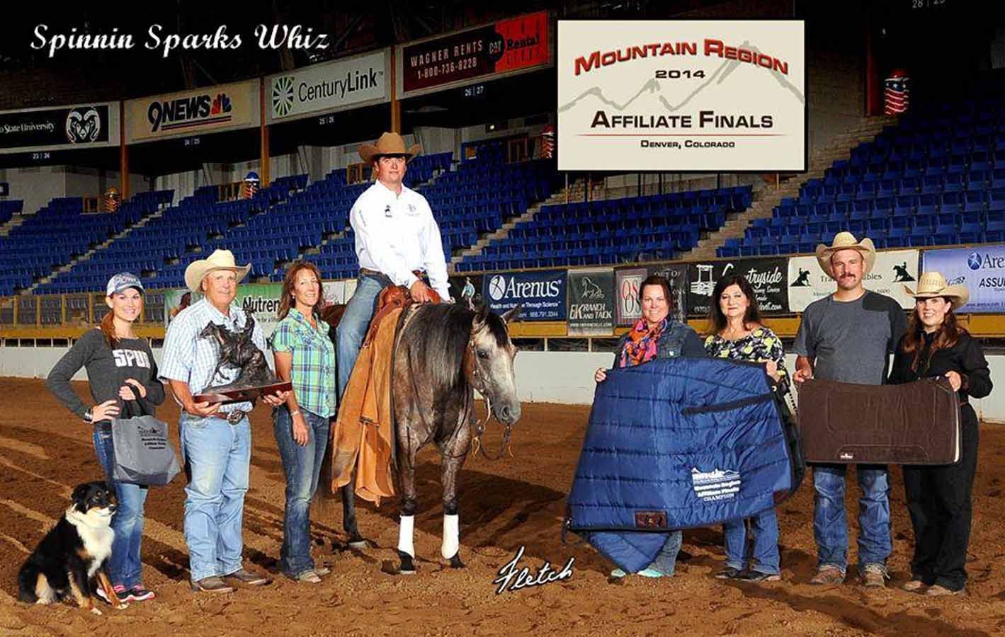 Award ceremony in an indoor arena with a rider on a horse, several people, and a dog near sponsor banners.