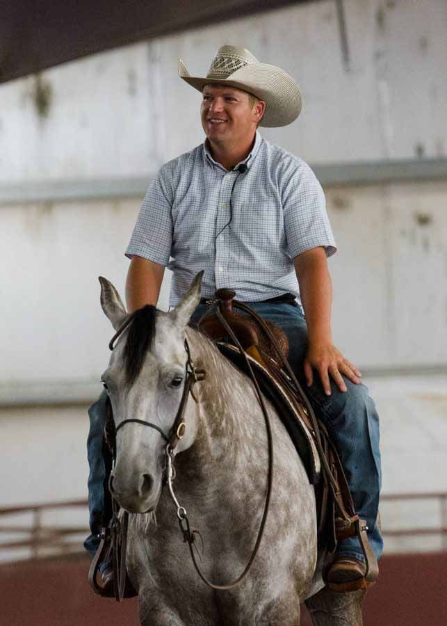Man in a cowboy hat smiling while riding a gray horse indoors
