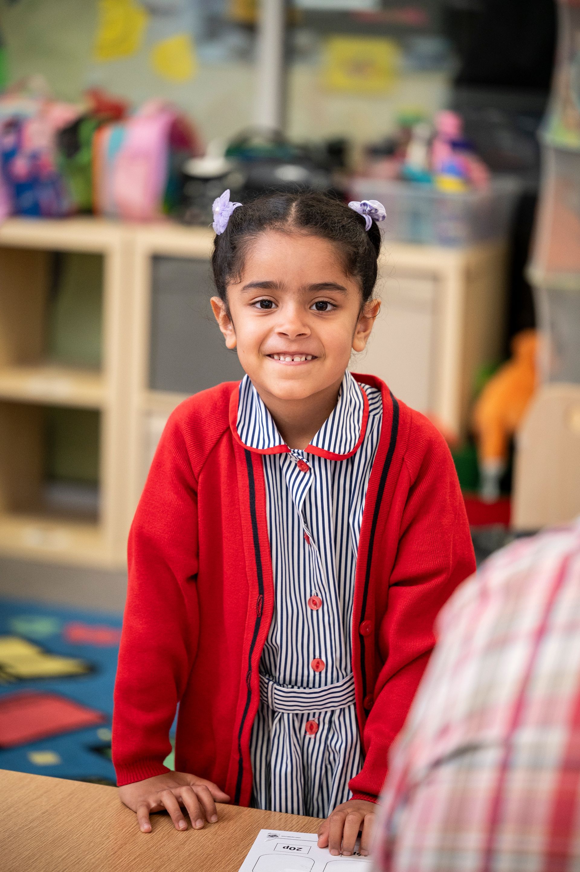 A little girl in a red sweater is sitting at a table in a classroom.