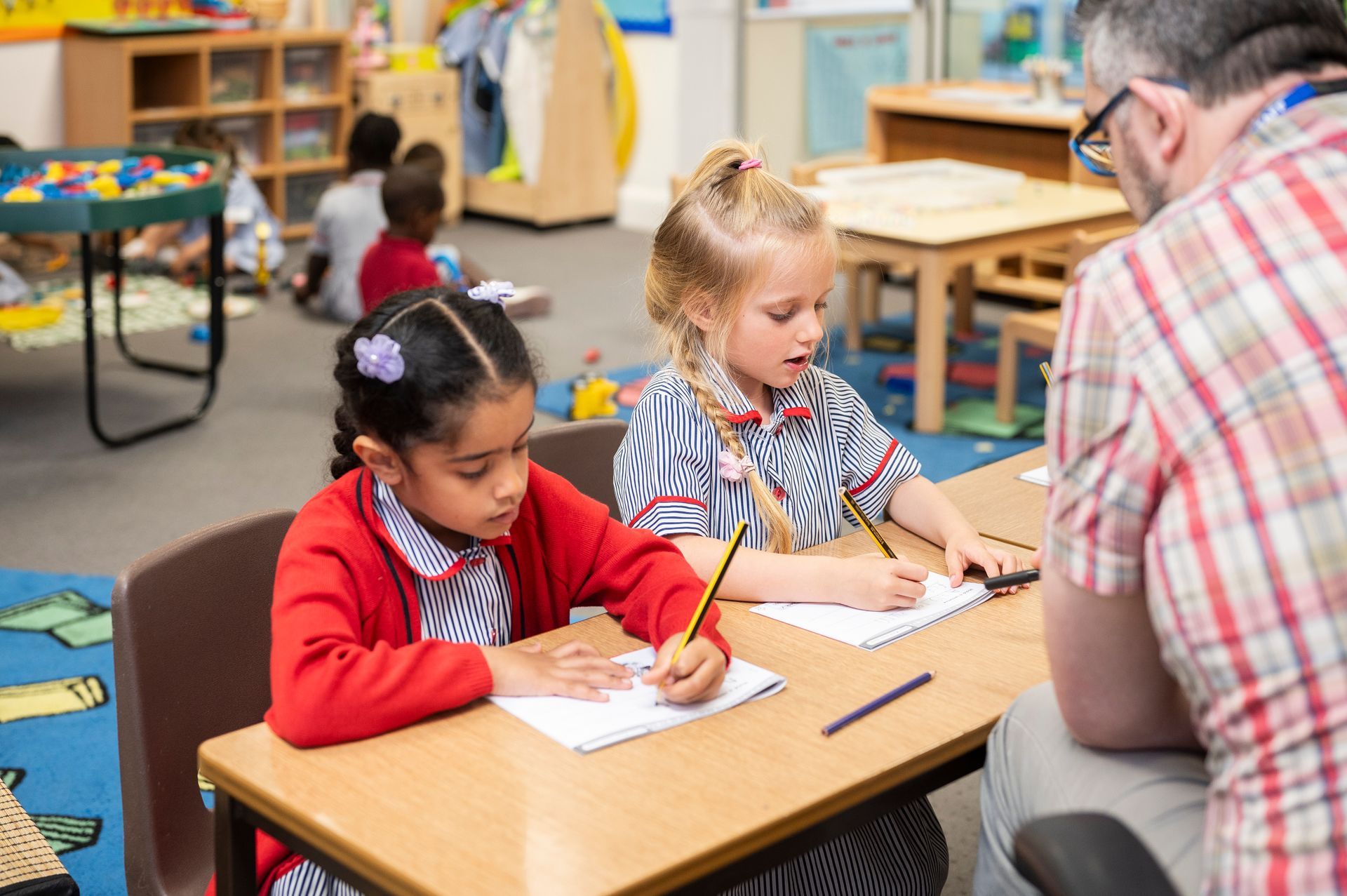 Two little girls are sitting at a desk in a classroom with a man.