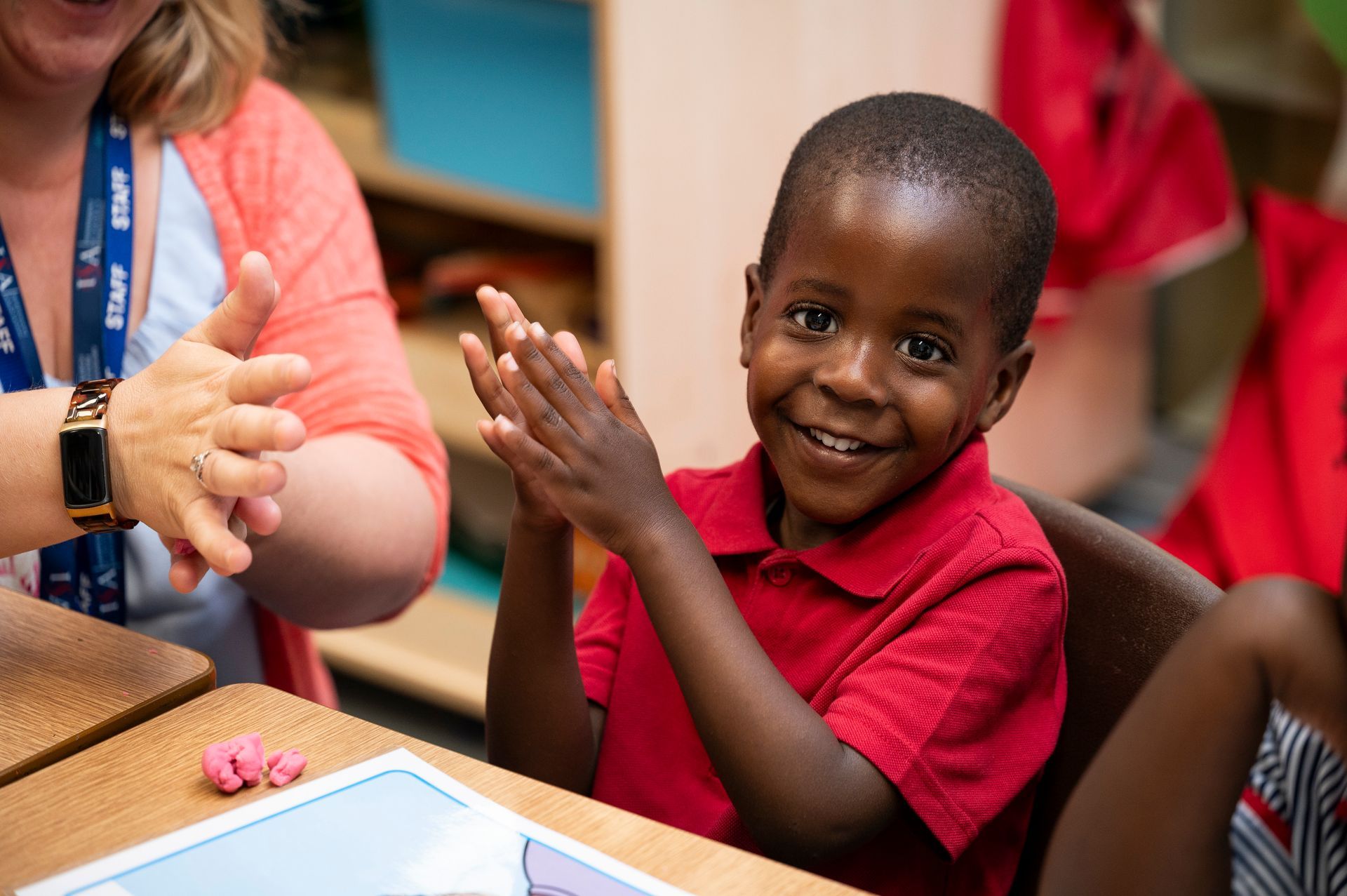 A young boy in a red shirt is clapping his hands in a classroom.