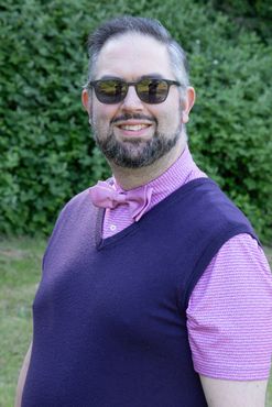 A man in a blue shirt is smiling for the camera in front of a bush.