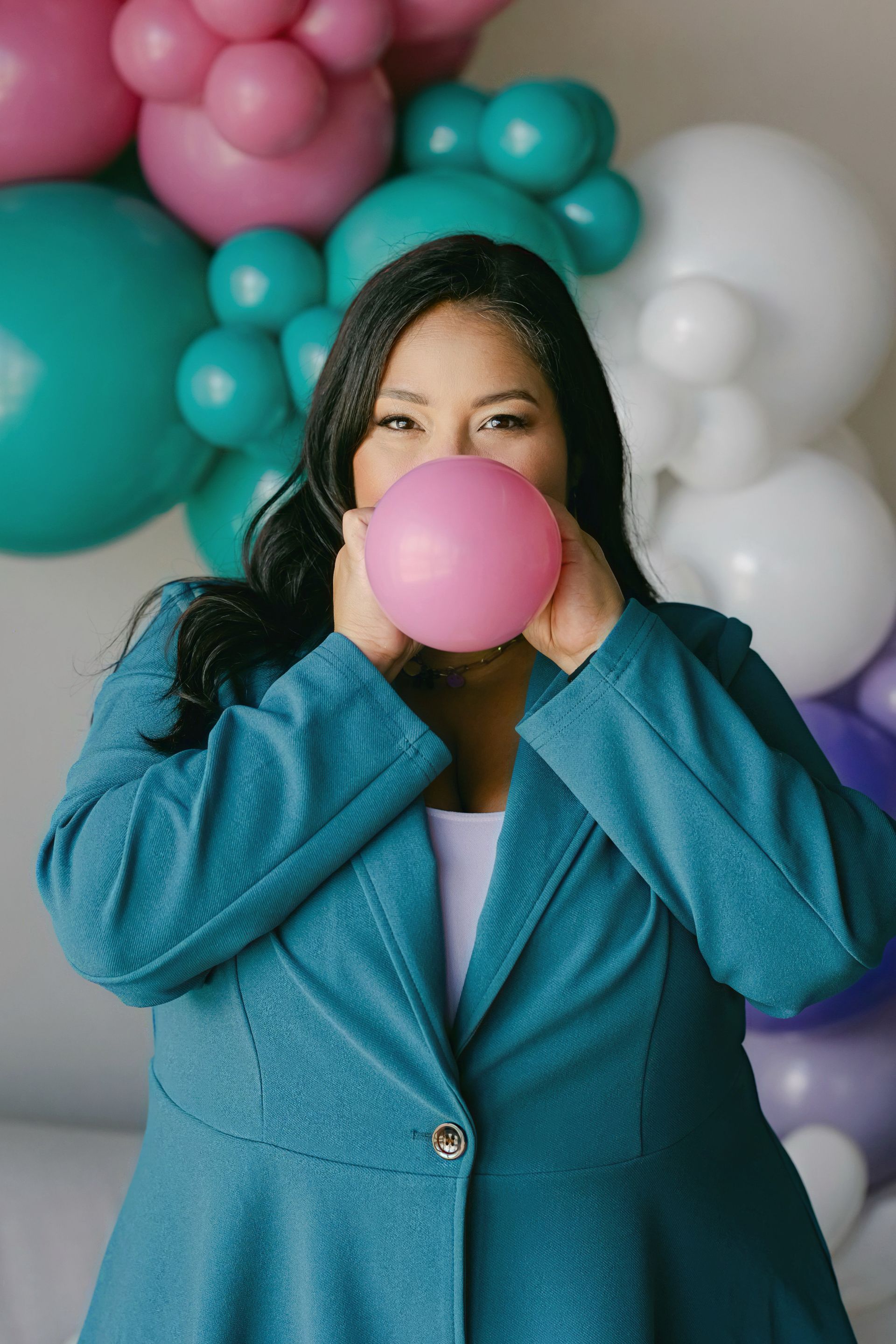 Person in a teal jacket blowing a pink bubble gum bubble in front of pastel balloons