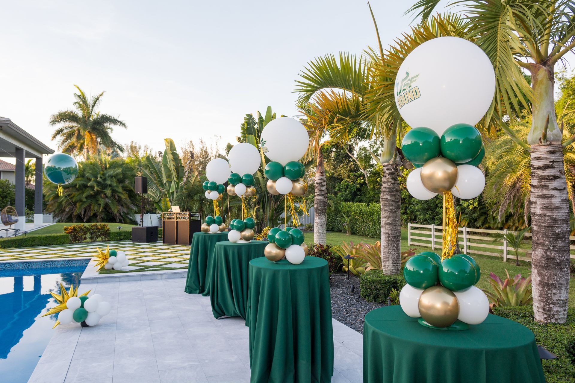 Outdoor poolside event decor with green, white, and gold balloon centerpieces along draped tables