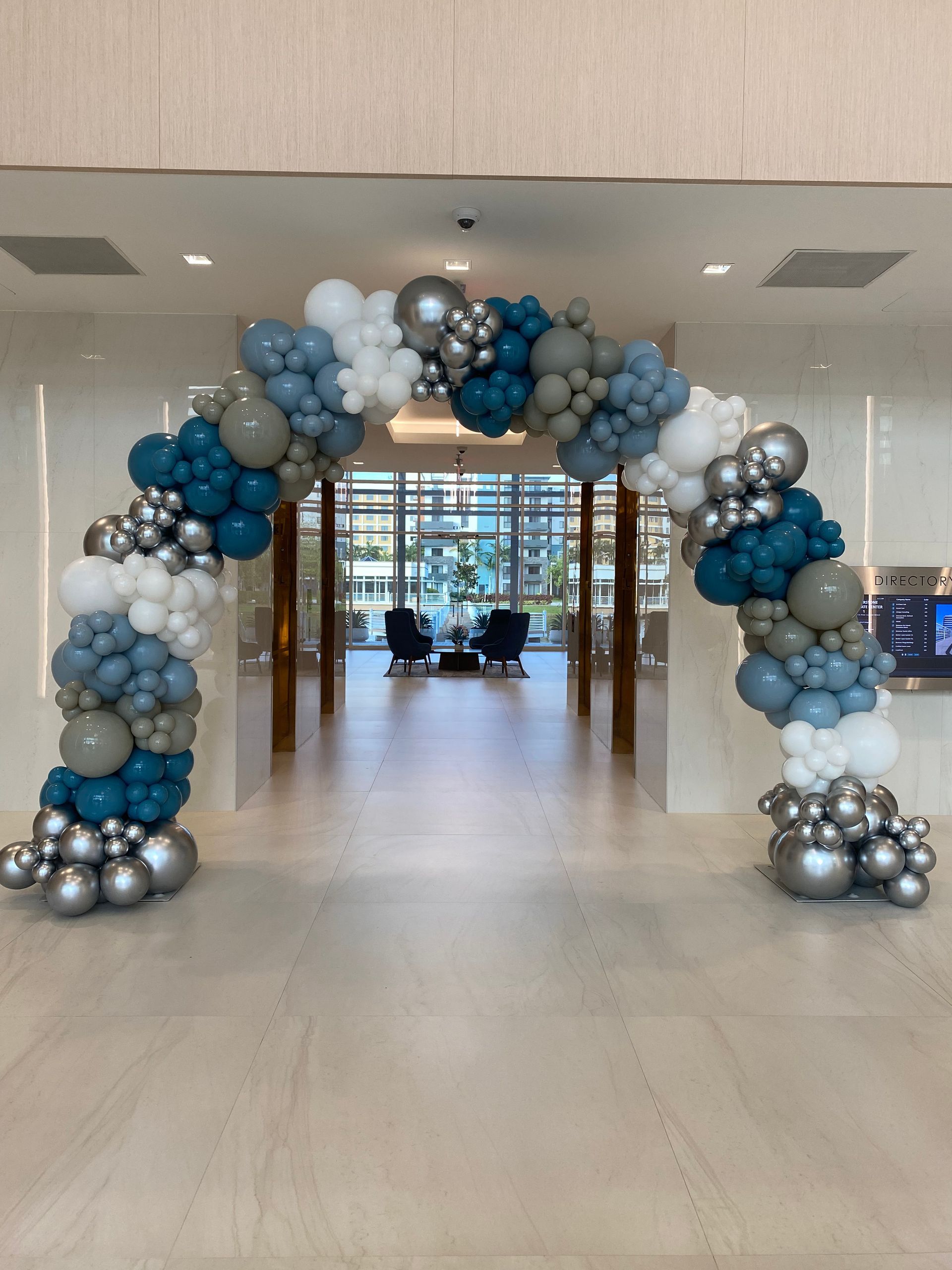 Blue, gray, and silver balloon arch framing an event hallway with windows beyond