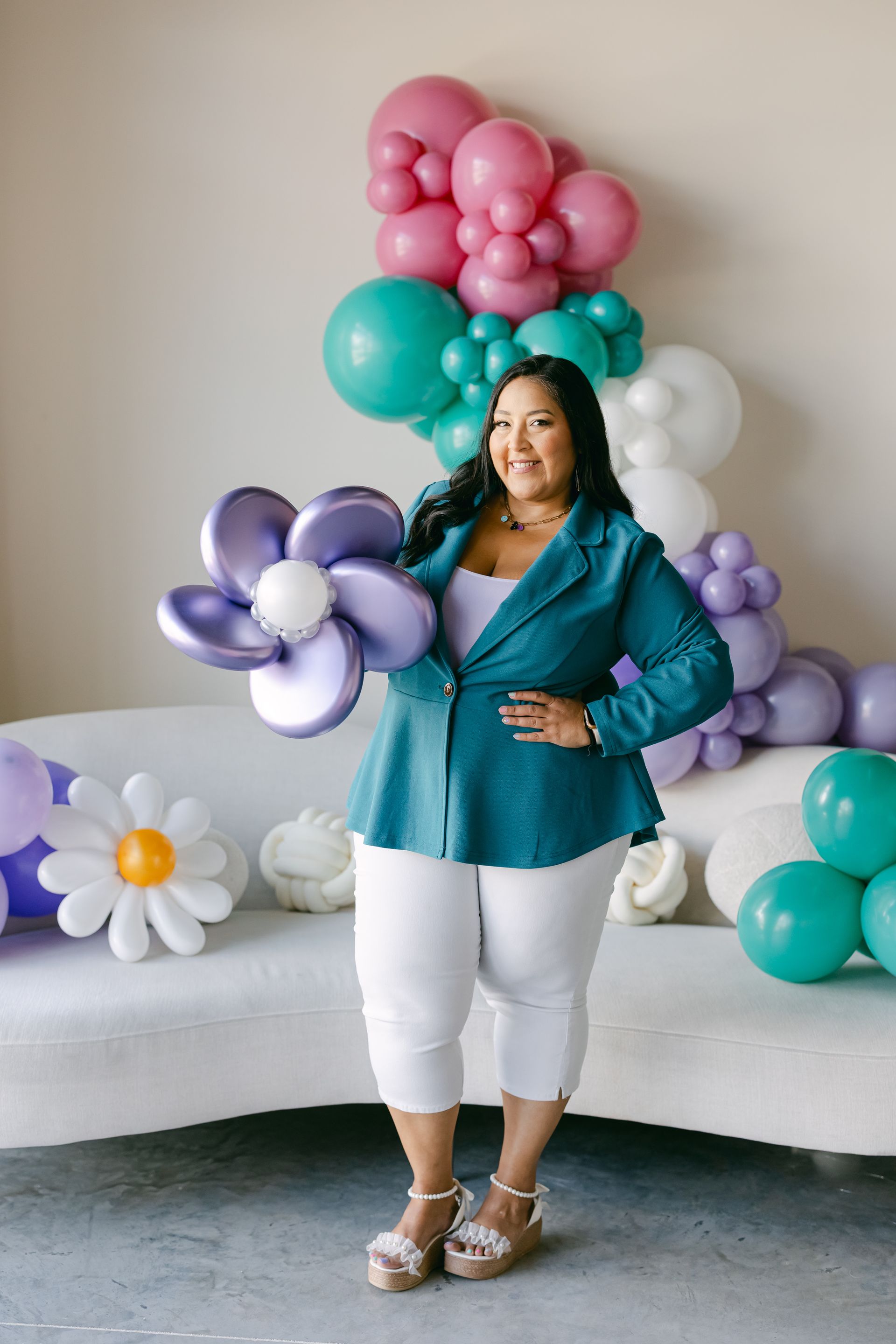 Woman in teal blouse and white pants posing with pastel balloon flowers and a white couch backdrop