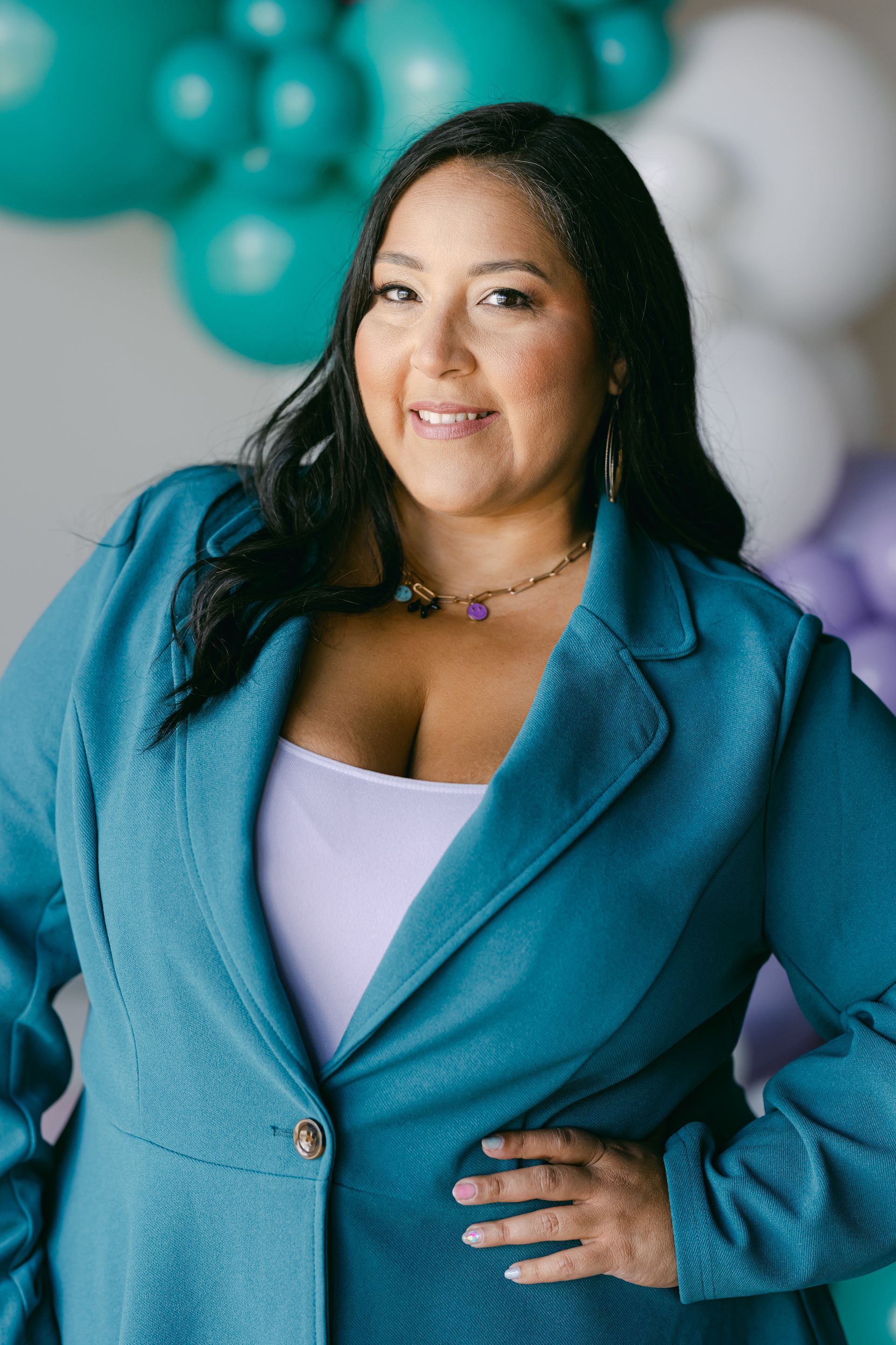 Woman in a teal blazer poses with hand on hip against a soft balloon backdrop.